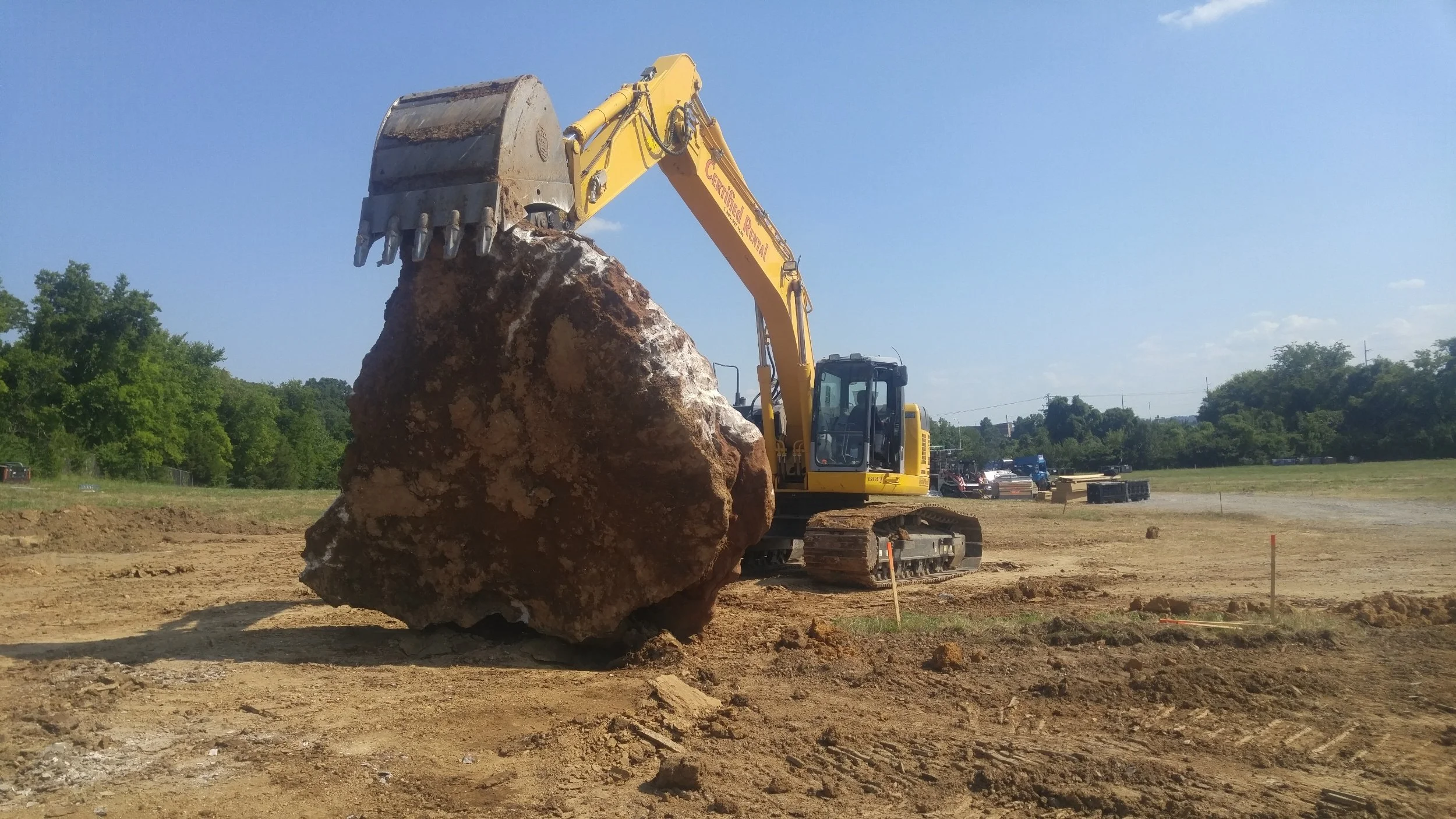 A yellow excavator lifting a large dirt and rock boulder at a construction site under a clear blue sky.