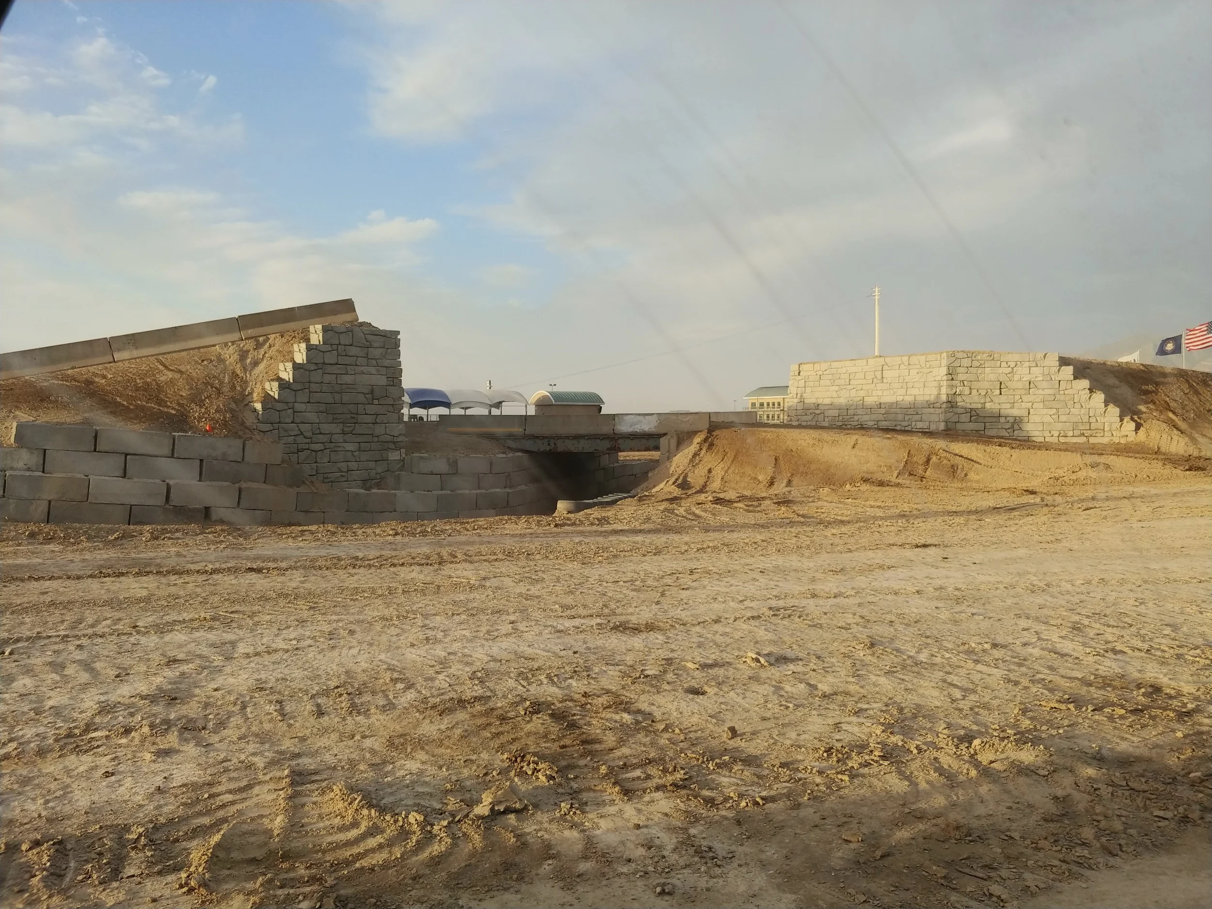 A construction site with unfinished brick walls, dirt ground, and a partly cloudy sky. There are flags, buildings, and training structures in the background.