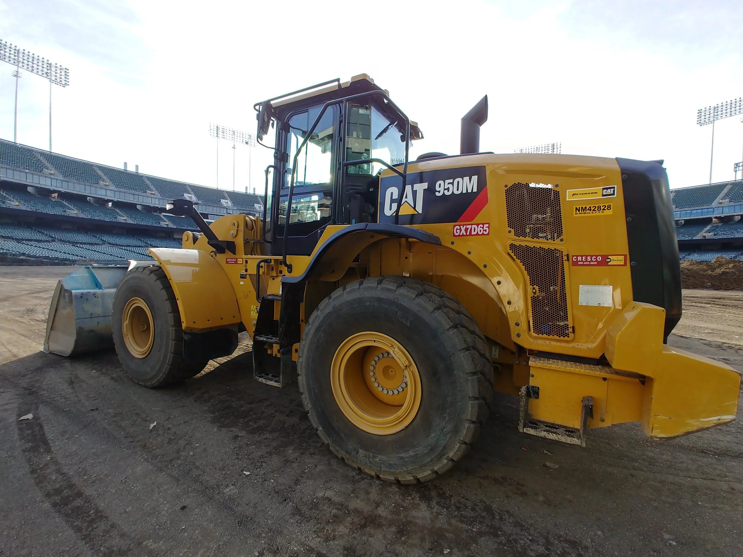 A yellow Caterpillar 950M wheel loader on a dirt surface, with a stadium with empty seats and tall floodlights in the background.