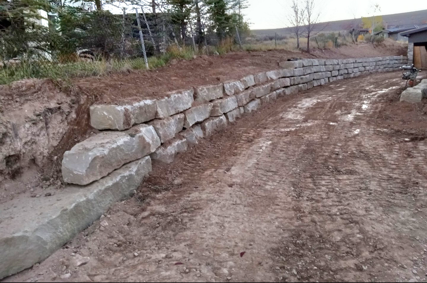 A beautifully stacked rock and stone wall which is curved and under construction with large stones and boulders stacked in rows, dirt and tracks in the foreground depicting a construction site with a 30 degree slope upwards