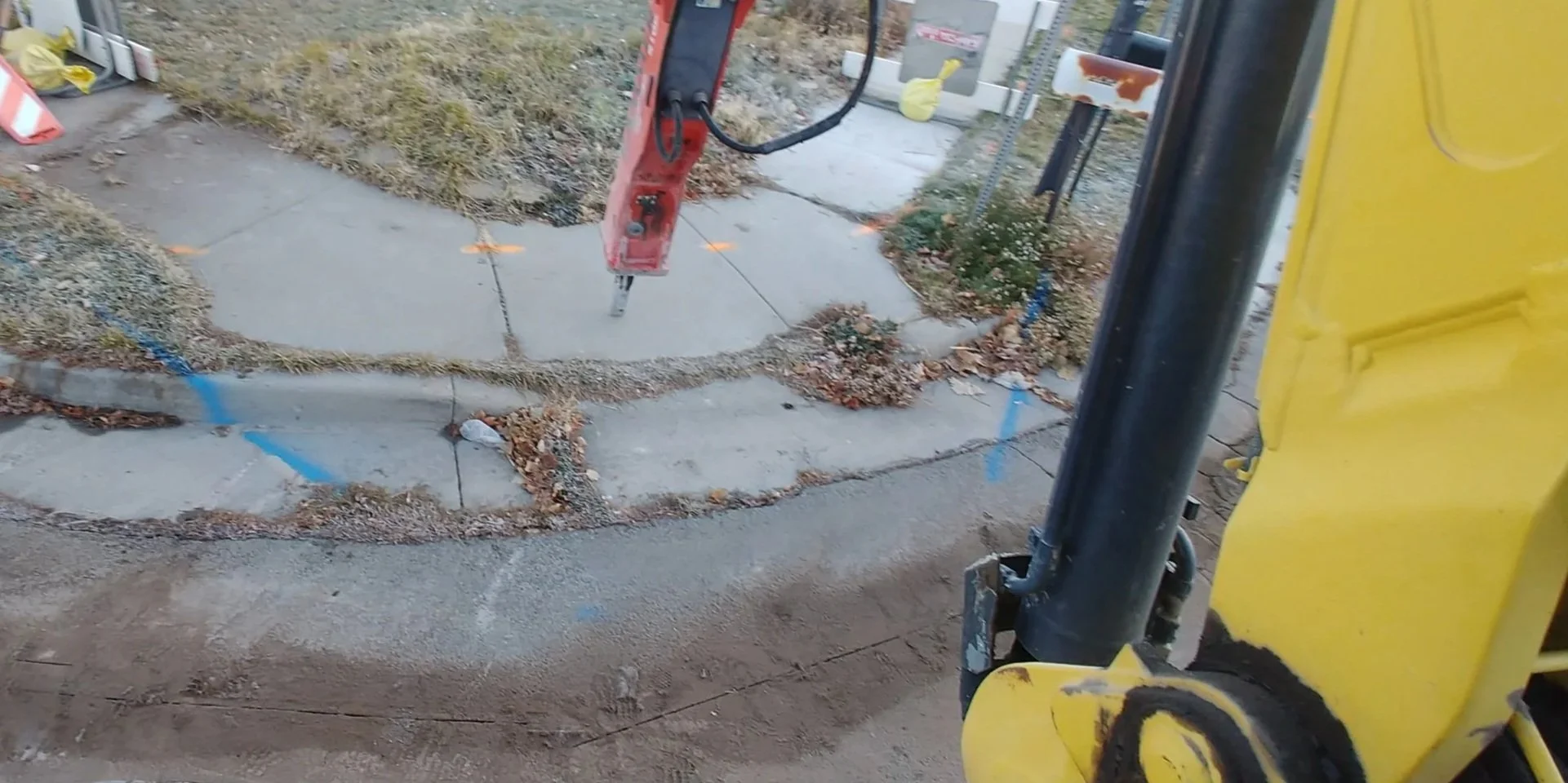 Construction site with sidewalk being broken with hydraulic hammer to remove and be repaired using heavy machinery, yellow equipment, and marked with blue spray paint, with leaves and dirt around.