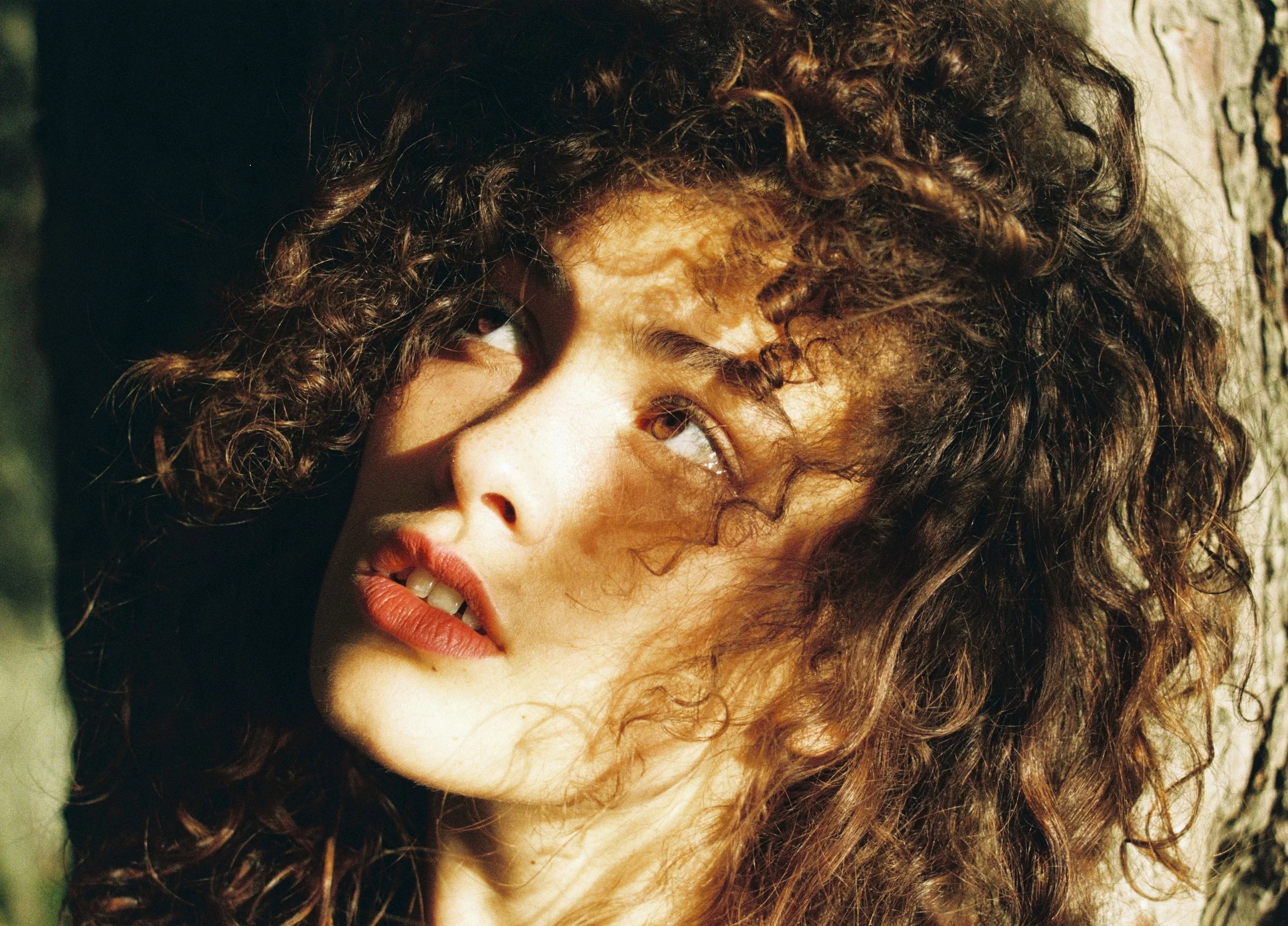 Close-up of a woman with curly brown hair leaning against a tree trunk, looking up and to her left, with sunlight illuminating her face.