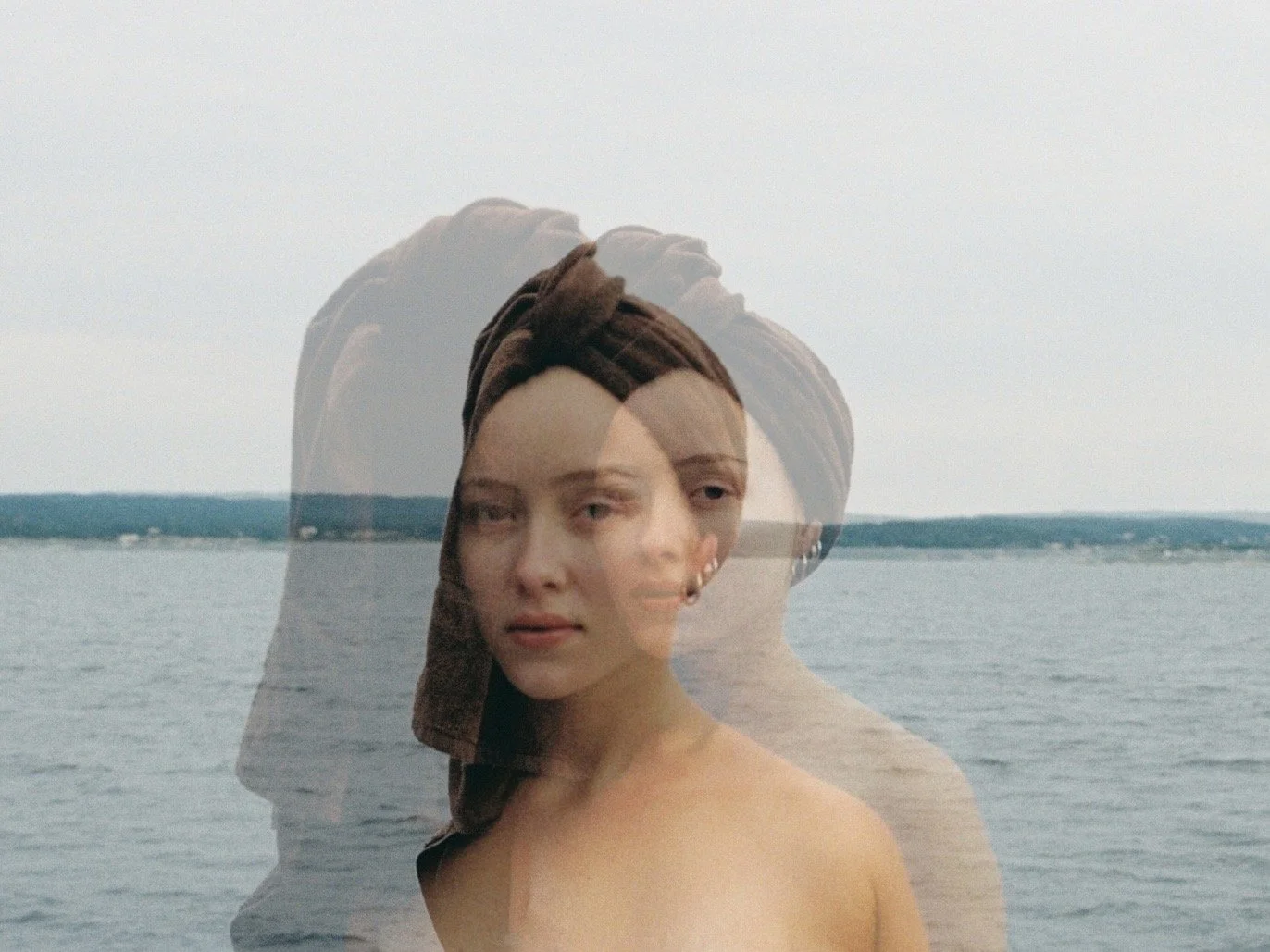 A young woman with a towel wrapped around her head standing by the water with a past overlay of her face, creating a double exposure effect.