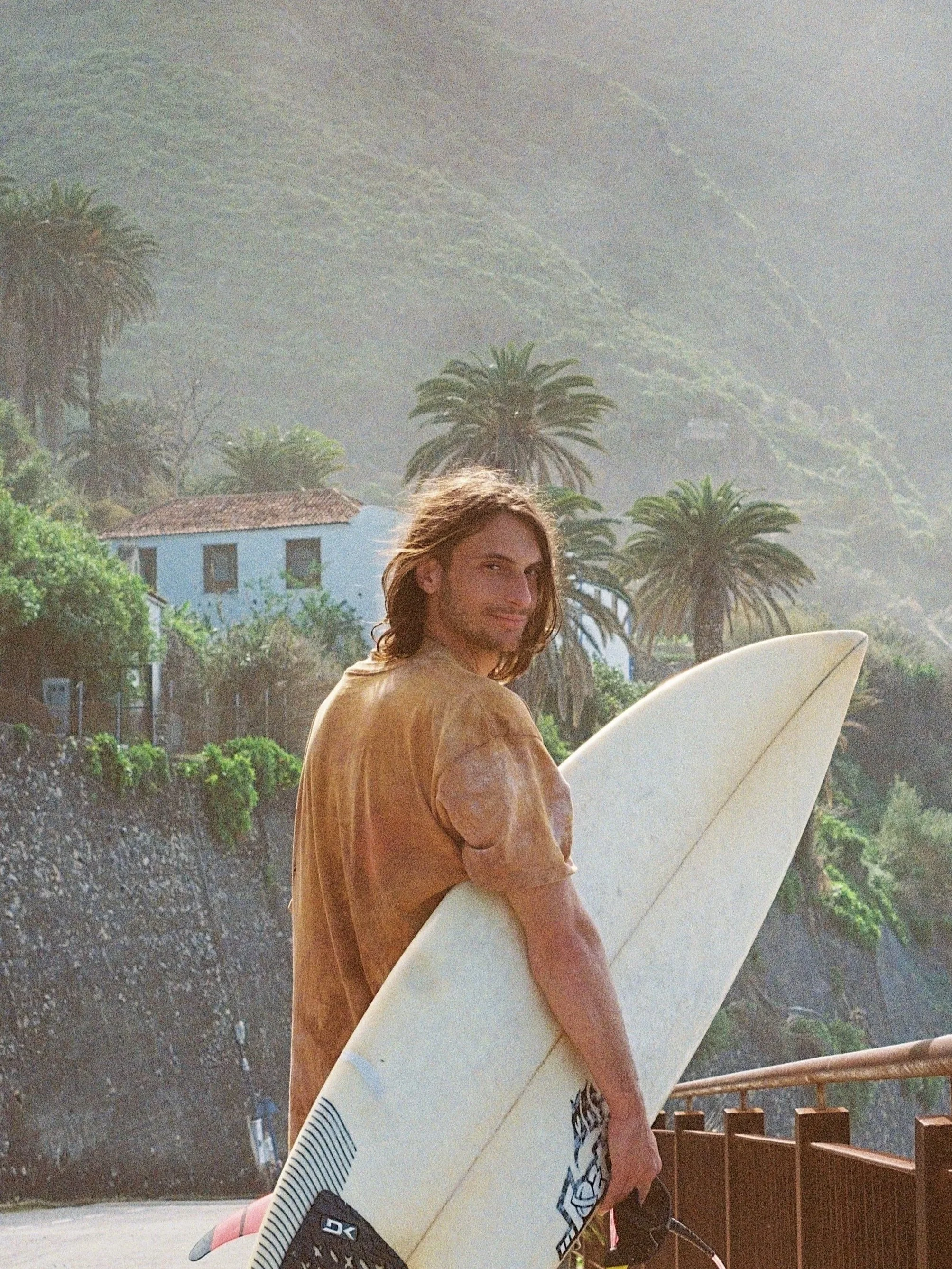 A man holding a surfboard while standing outdoors with palm trees and hillside in the background.