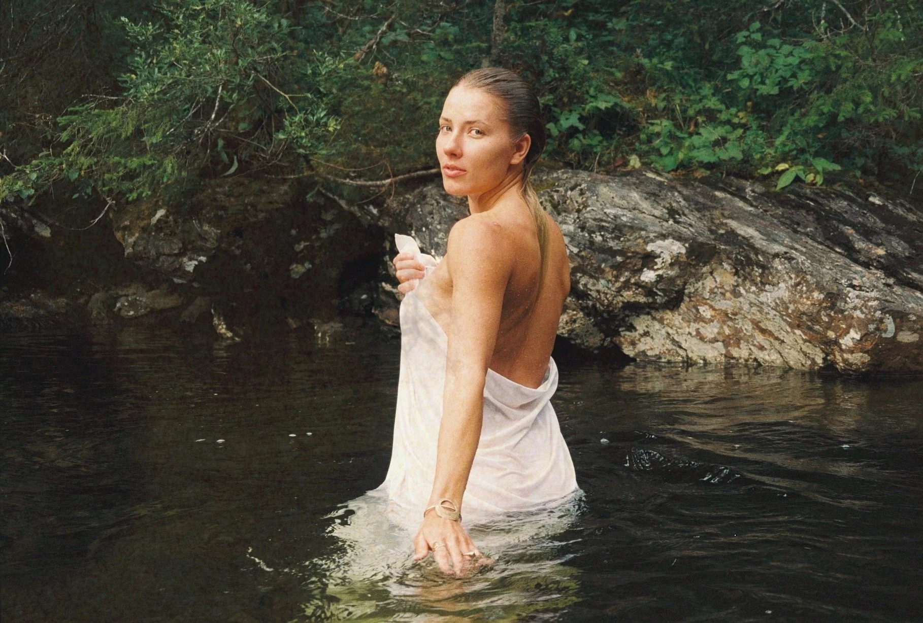 A woman with wet hair standing in a river, holding a white cloth around her waist, with rocks and green foliage in the background.