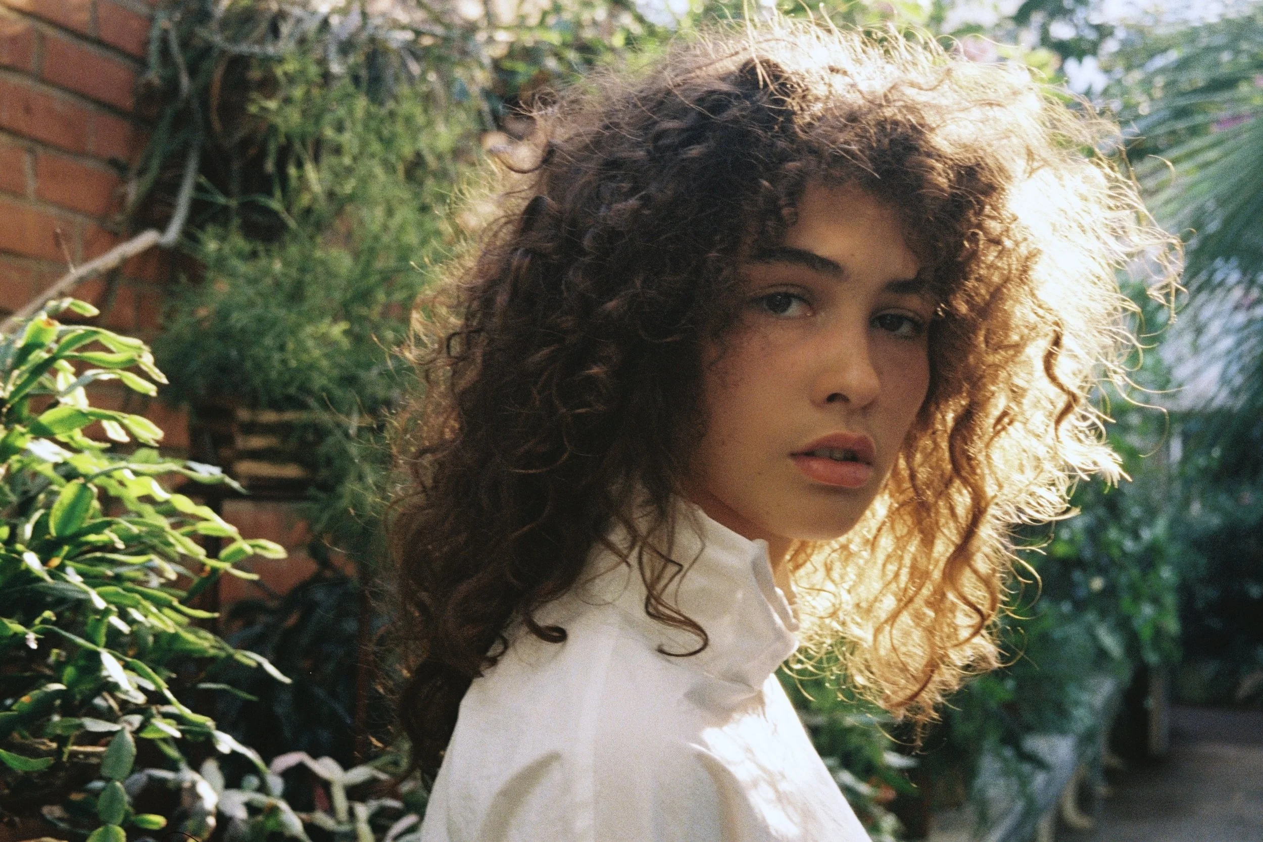 A young woman with curly hair and light skin looking at the camera. She is outdoors with greenery and a brick wall in the background, illuminated by sunlight.