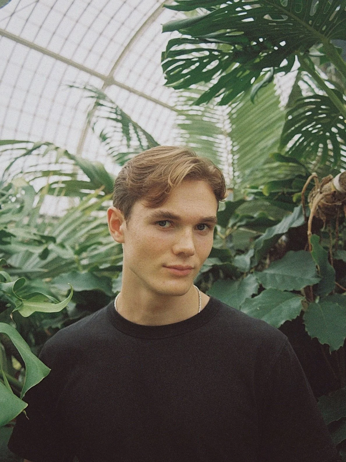 A young man with blonde hair and a black shirt poses in front of tropical plants inside a greenhouse.