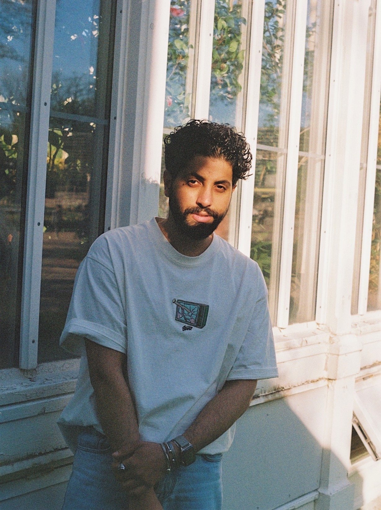A young man with curly hair and a beard standing outside near a large window, wearing a white t-shirt with a colorful graphic of a TV and some bracelets on his left wrist.