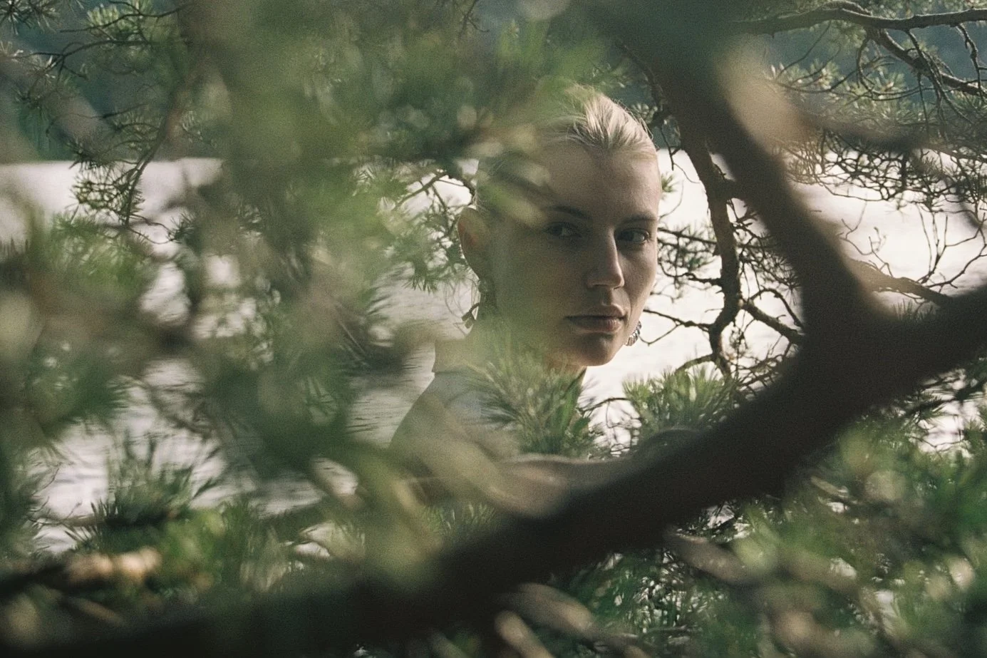 A woman with blonde hair and hoop earrings looking through dense tree branches and leaves.