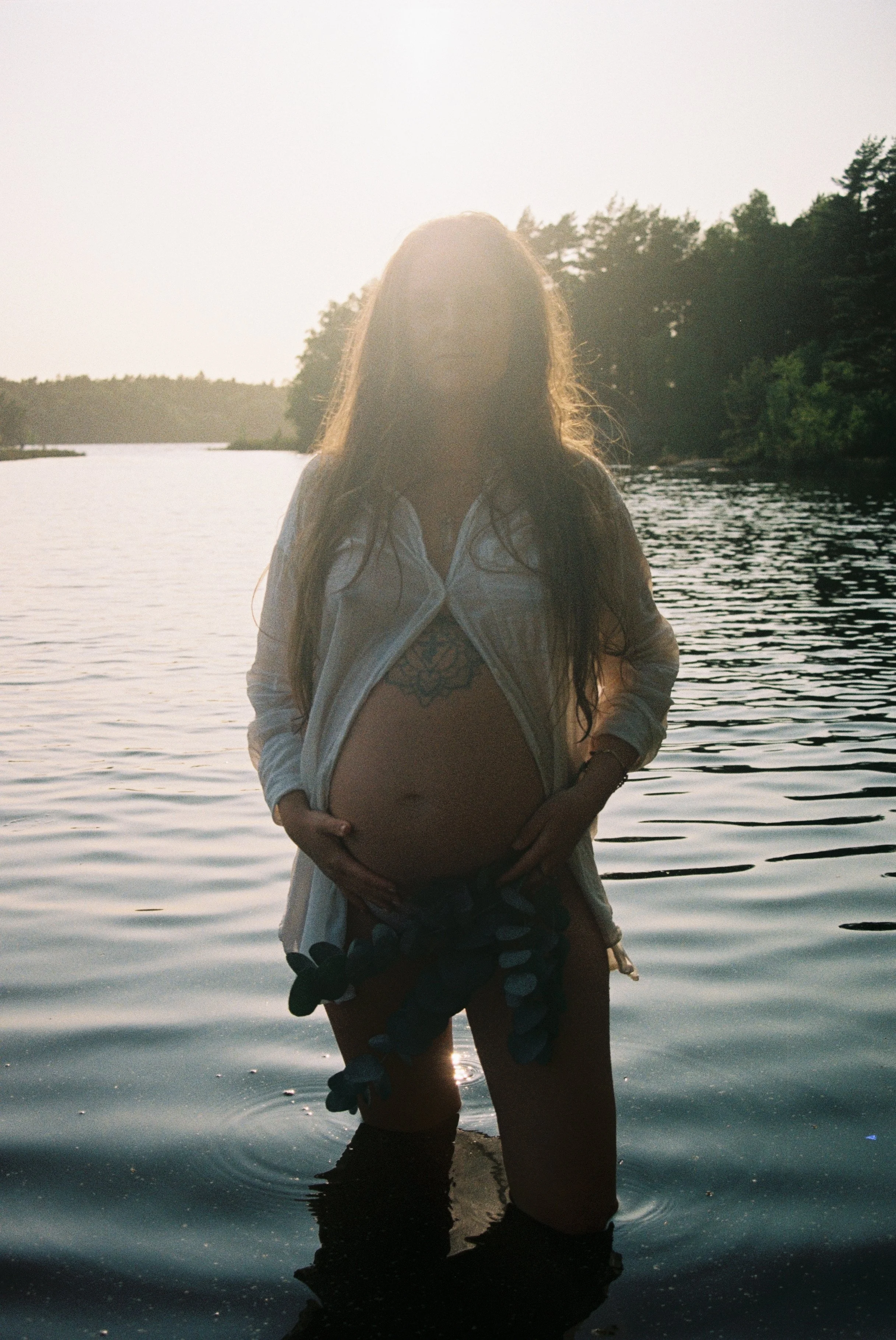 A pregnant woman standing in a body of water with her hands on her belly, backlit by the sun, with trees in the background.