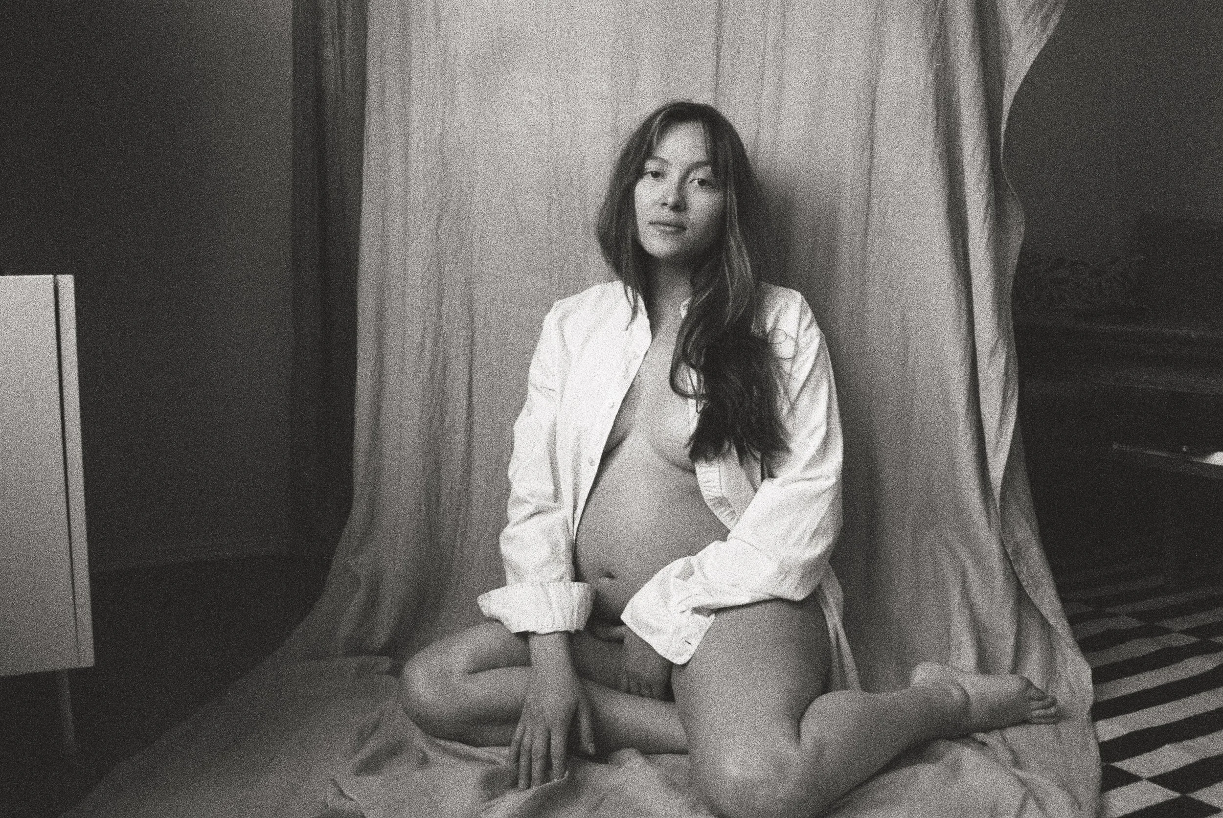 A black and white photo of a woman with long hair sitting on a bed, partially covered by a white shirt, in a room with curtains and a checkered floor.