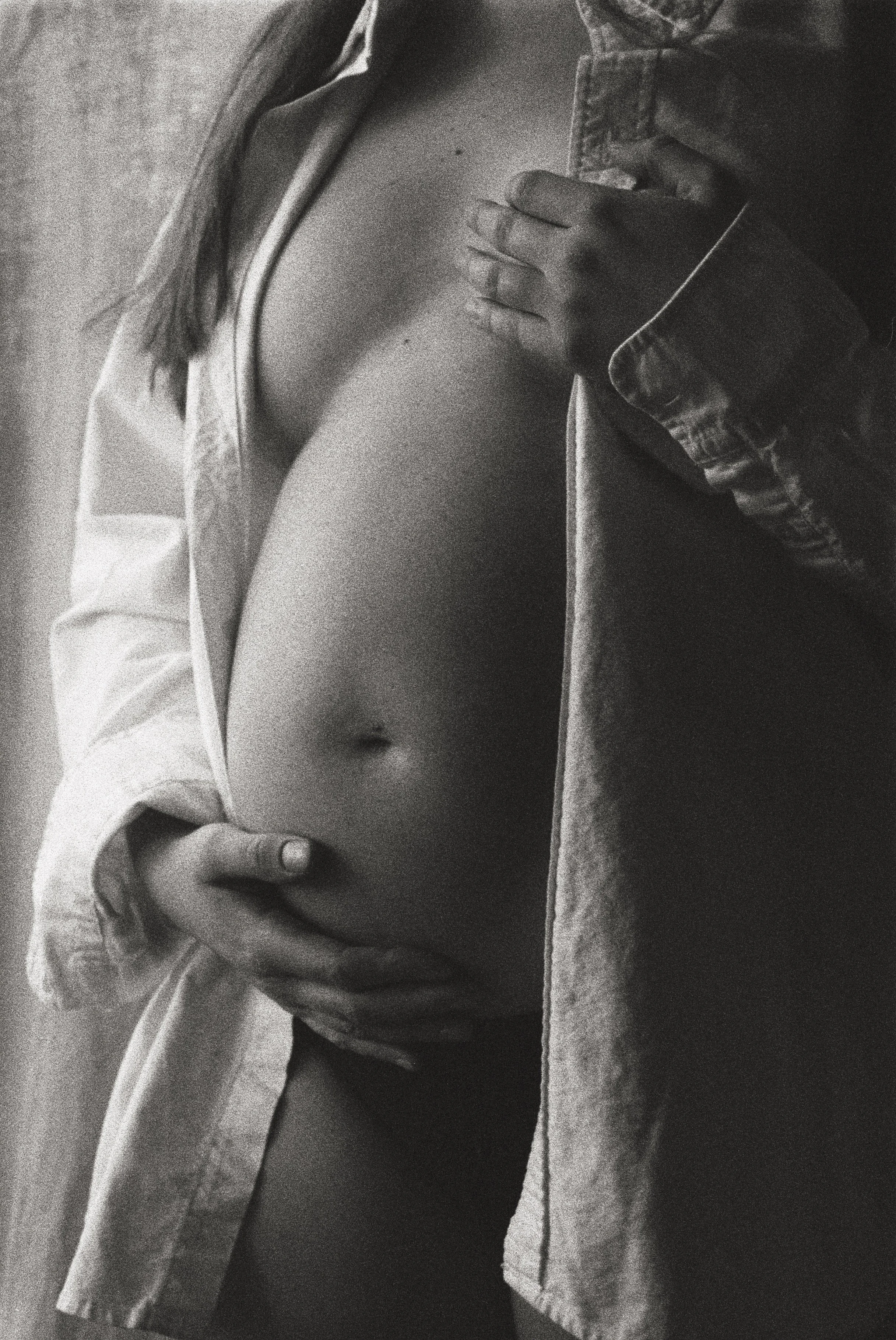 A black and white photo of a pregnant woman showing her bare belly, with one hand resting on her stomach and the other hand near her chest. She is wearing an unbuttoned denim jacket and a partially visible shirt.