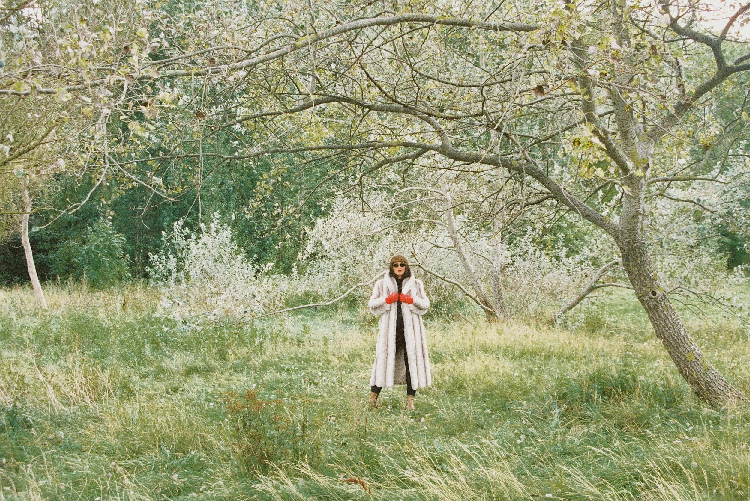 A woman standing in a grassy field under a tree with spreading branches, wearing a long coat, sunglasses, and red gloves.