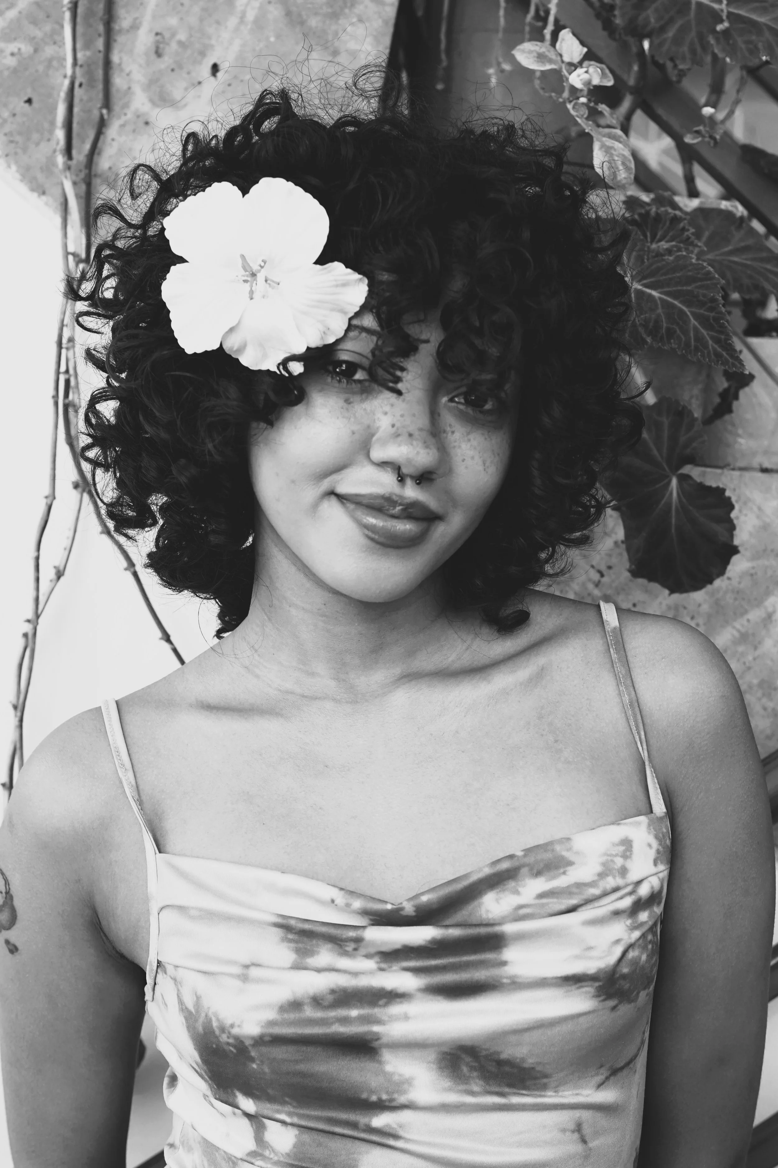 Creative hair salon apprentice at Gem House Salon in Greenpoint, Brooklyn, with natural curly hair and freckles, wearing a tie-dye dress and septum piercing, standing among plants, reflecting an inclusive, artistic salon team for emerging stylists.