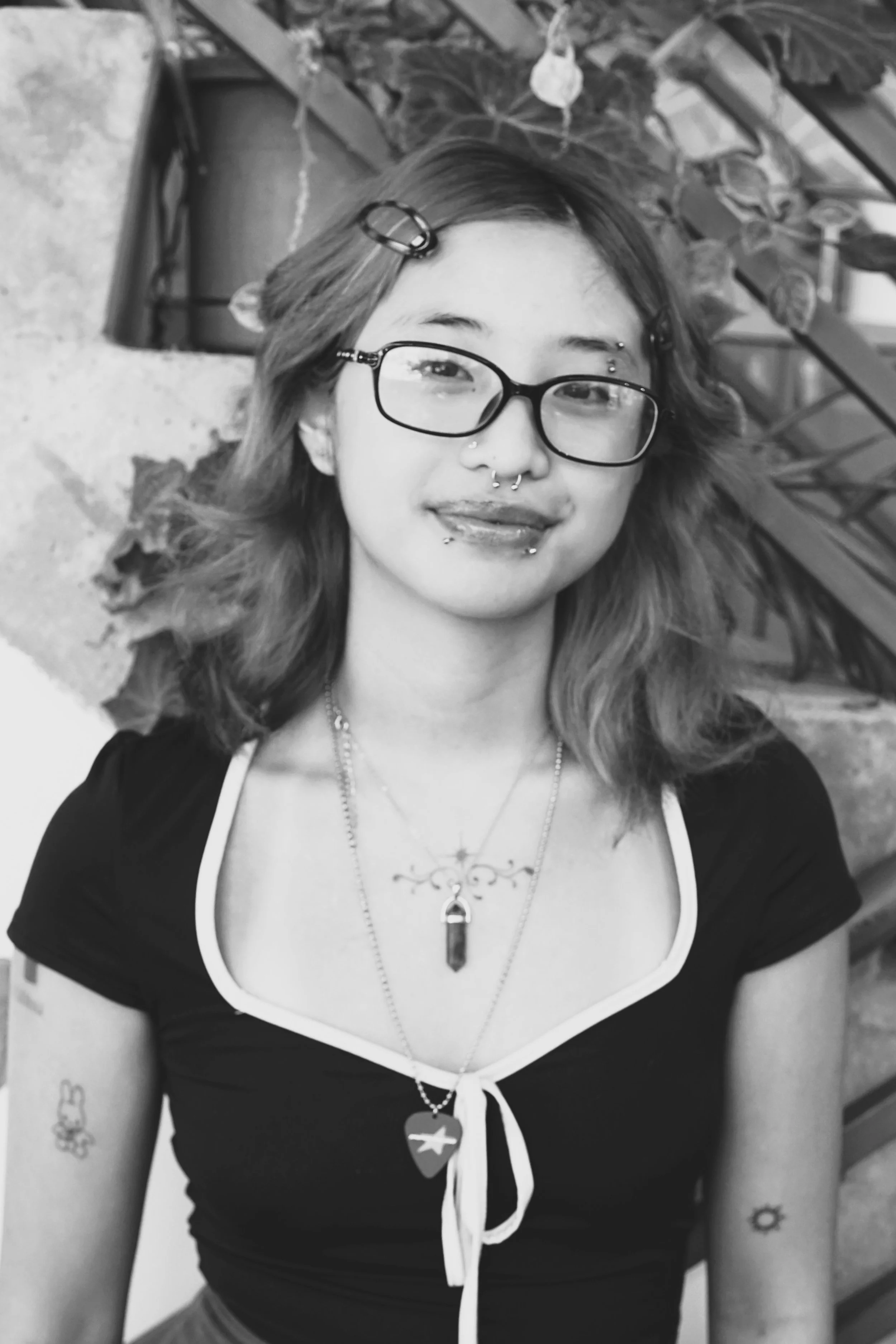 Hair salon apprentice at Gem House Salon in Greenpoint, Brooklyn, wearing glasses and hair clips, standing outdoors among plants and a brick wall, representing a supportive, creative salon team environment for aspiring stylists.