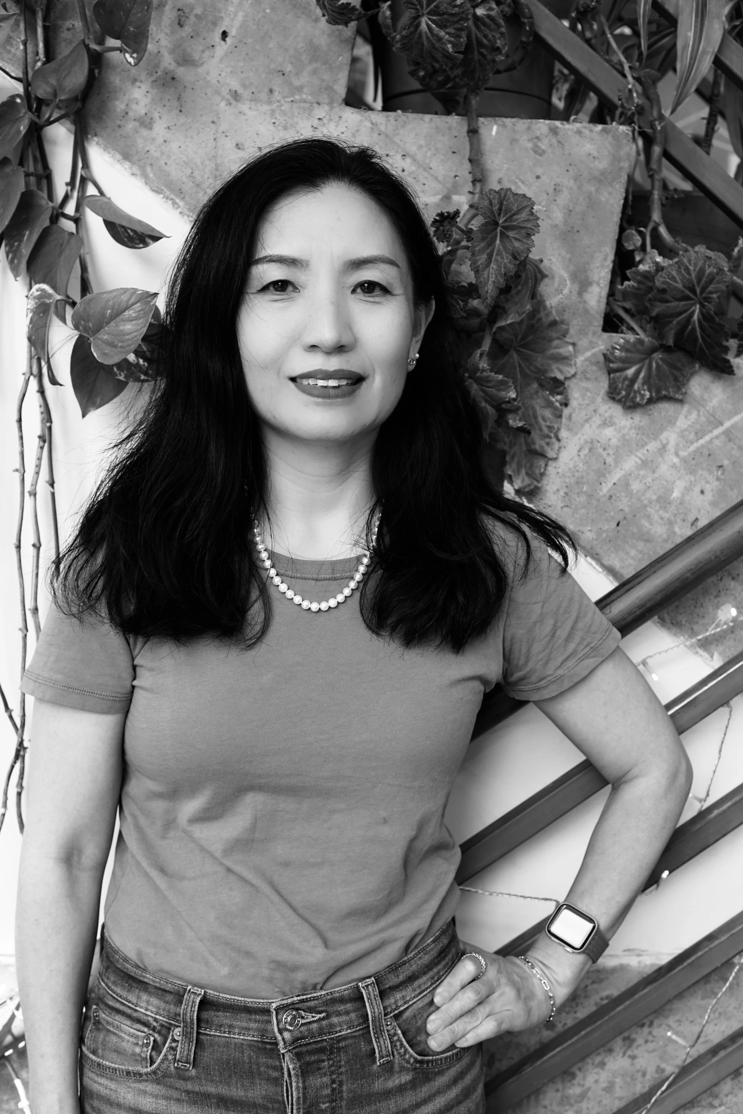 Lived-in blonde and bronde hair color specialist at Gem House Salon in Greenpoint, Brooklyn, smiling with long wavy hair and arm tattoos in a black and white portrait seated near a staircase.