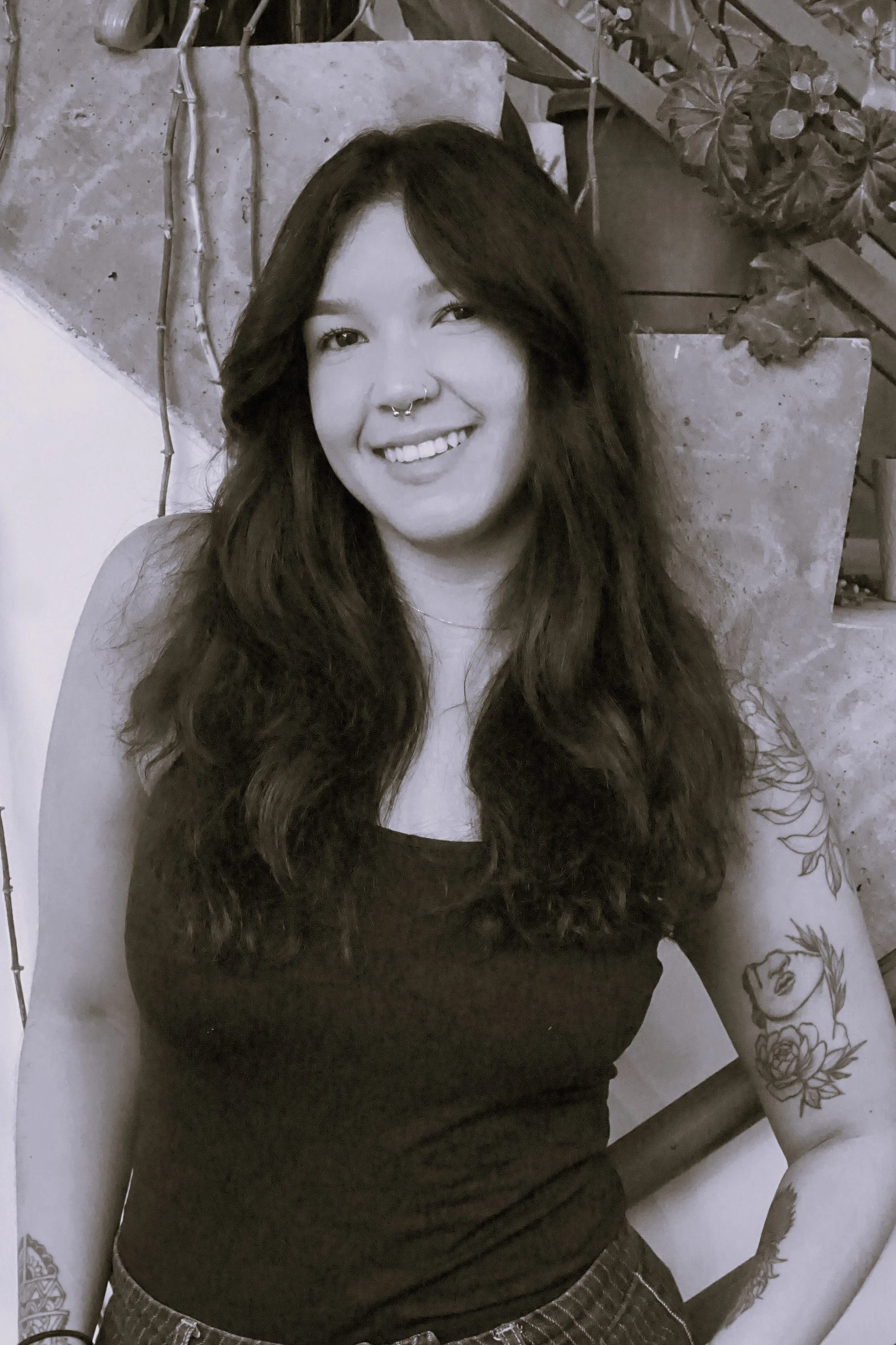 Lived-in blonde and bronde hair color specialist at Gem House Salon in Greenpoint, Brooklyn, smiling with long wavy hair and arm tattoos in a black and white portrait seated near a staircase.