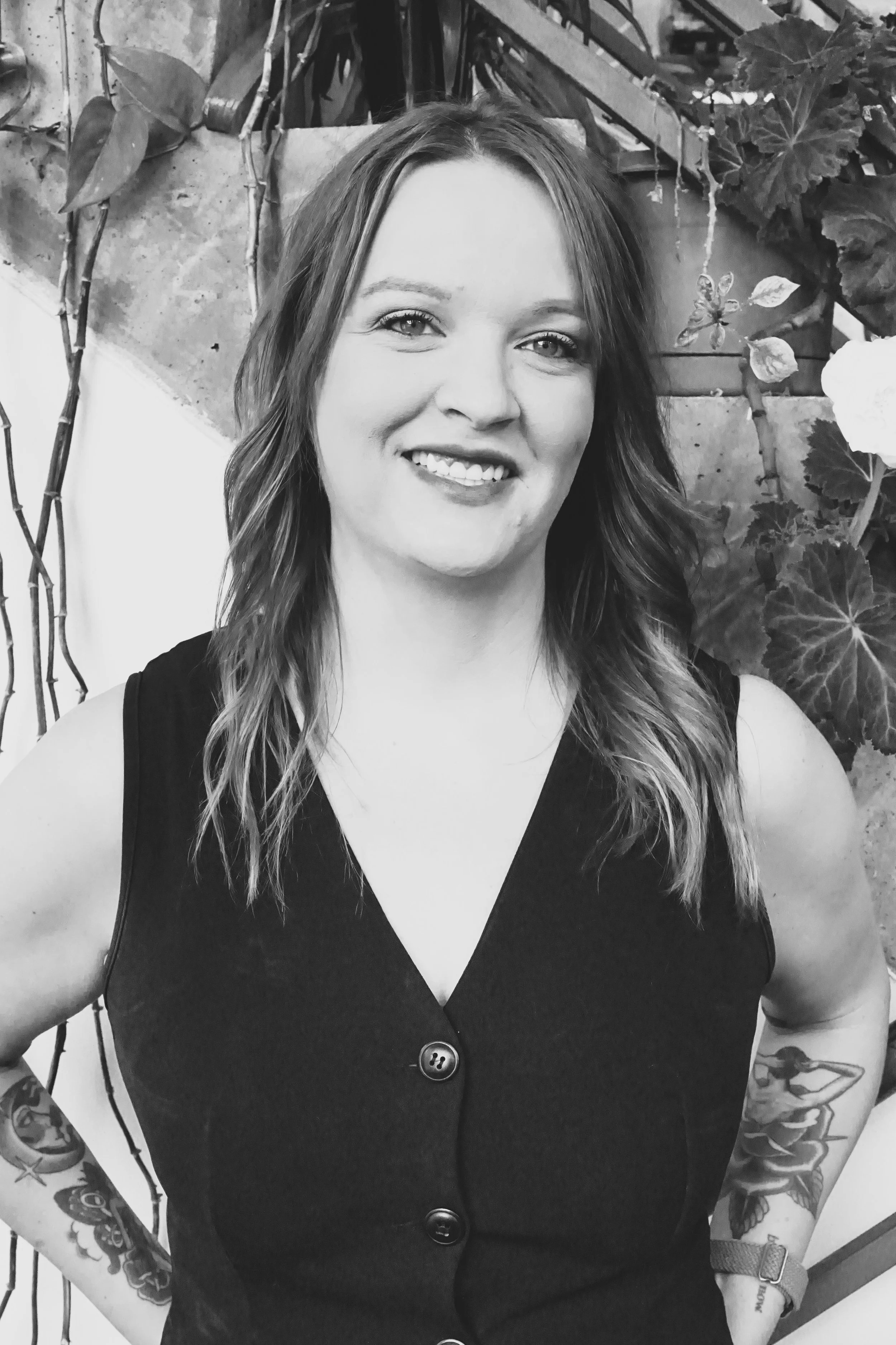 Razor haircut, keratin treatment, and wavy perm specialist at Gem House Salon in Greenpoint, Brooklyn, smiling with long wavy hair in a black and white portrait, standing near plants and a staircase.
