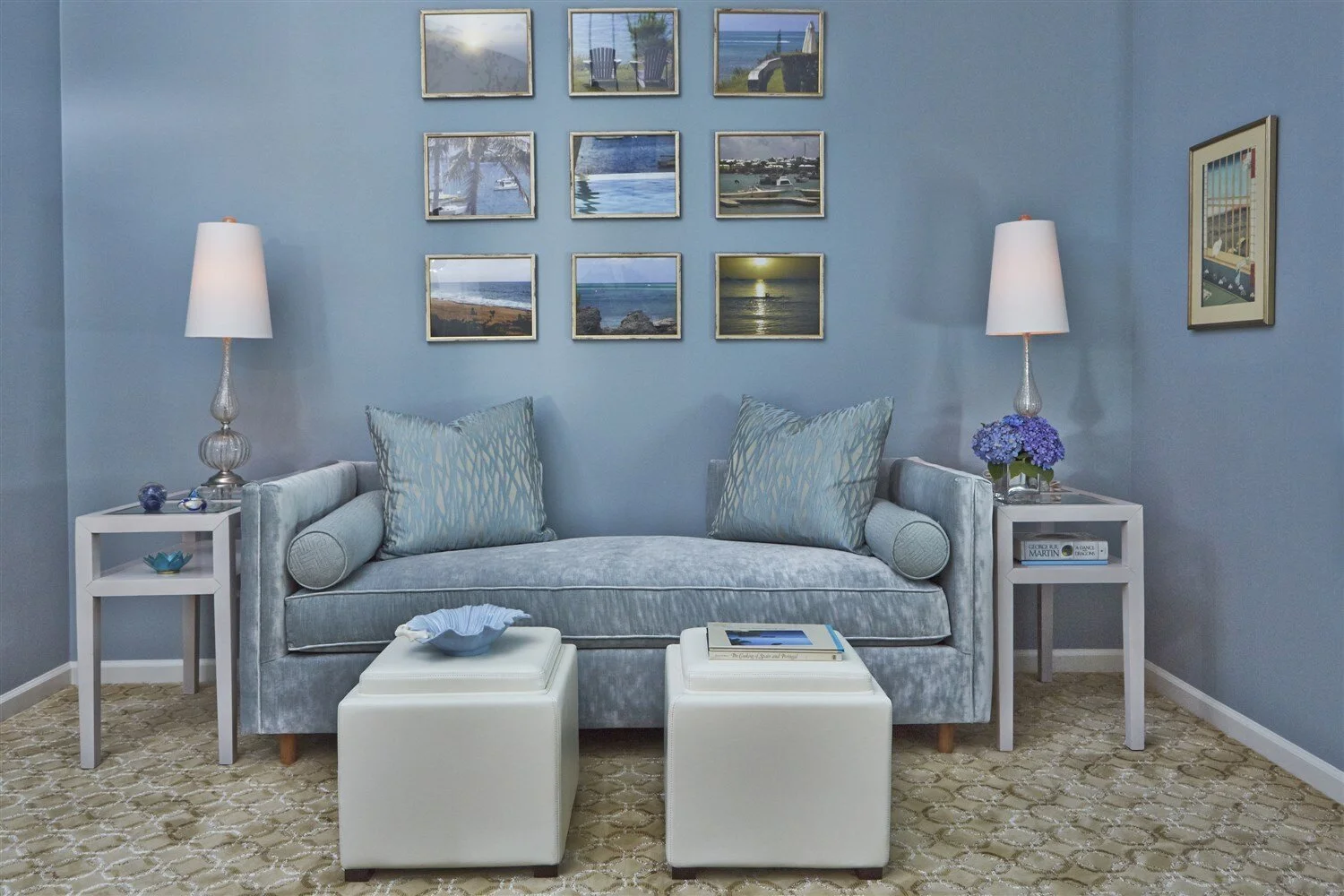 Coastal living room with light blue walls, a plush velvet sofa with patterned pillows, two white ottomans, and two white side tables with lamps, framed ocean photographs on the wall, and a beige patterned carpet.