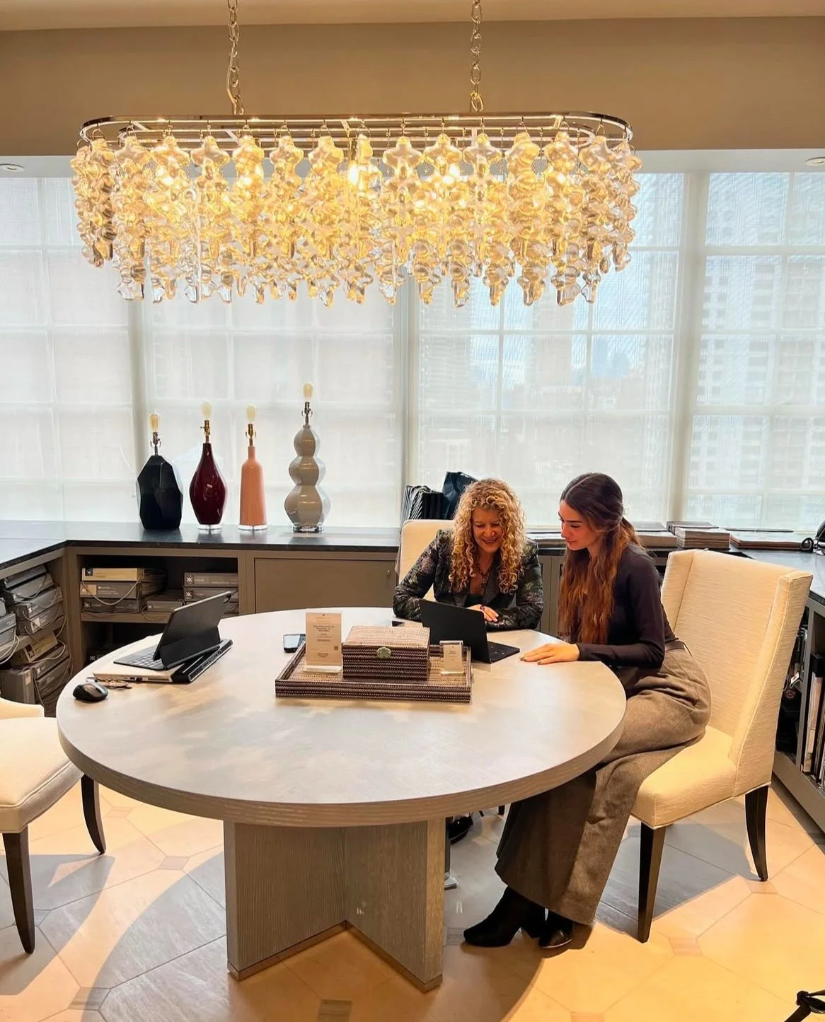 Two women sitting at a round table in a bright, modern office, looking at a laptop together, with a chandelier overhead and decorative vases in the background.