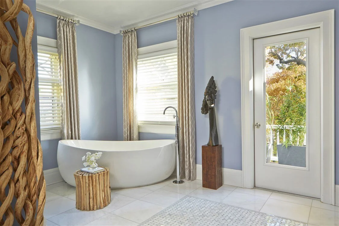 Modern bathroom with a freestanding bathtub near windows with patterned curtains, a wooden side table with flowers, an abstract sculpture on a pedestal, and a glass door leading outside to greenery.