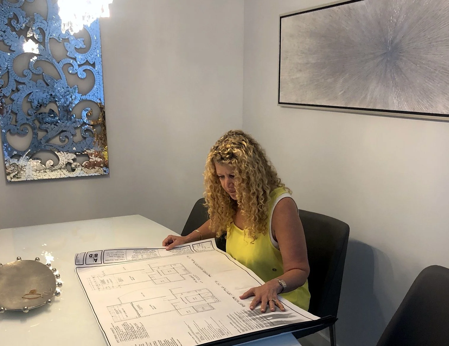 A woman with curly blonde hair wearing a yellow sleeveless top sits at a table, looking at an architect's blueprint. There is a decorative mirror on the left wall and a large abstract painting on the right wall.