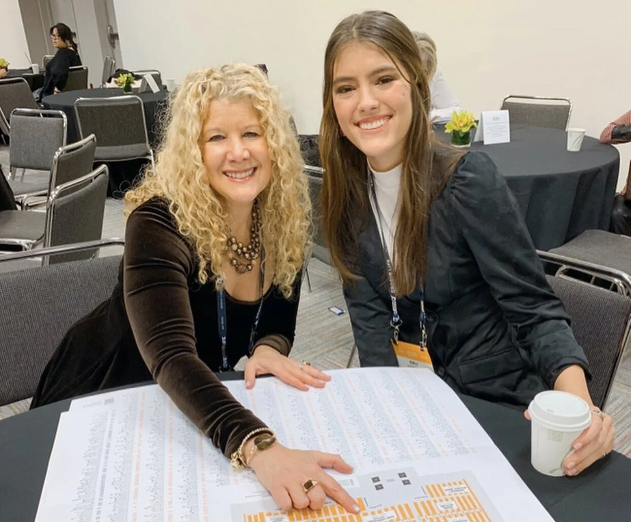 Two women sitting at a table, smiling at the camera, in a conference room. One woman has curly blonde hair, wearing a black top with a necklace, and the other woman has long straight brown hair, wearing a black jacket and holding a white coffee cup.