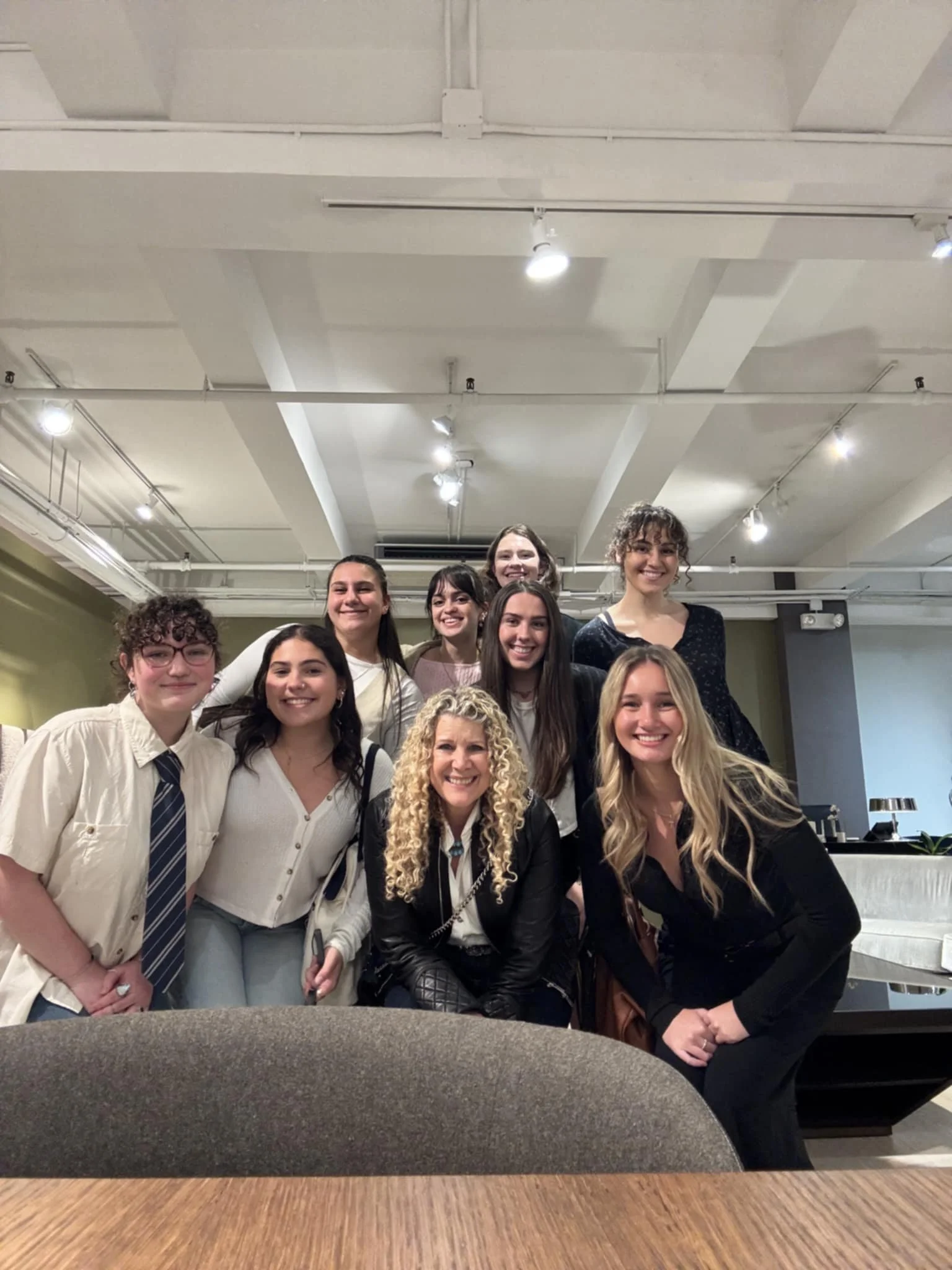 Group of nine women smiling and posing for a photo indoors, some standing and some kneeling, with a wooden table in the foreground and ceiling lights above.
