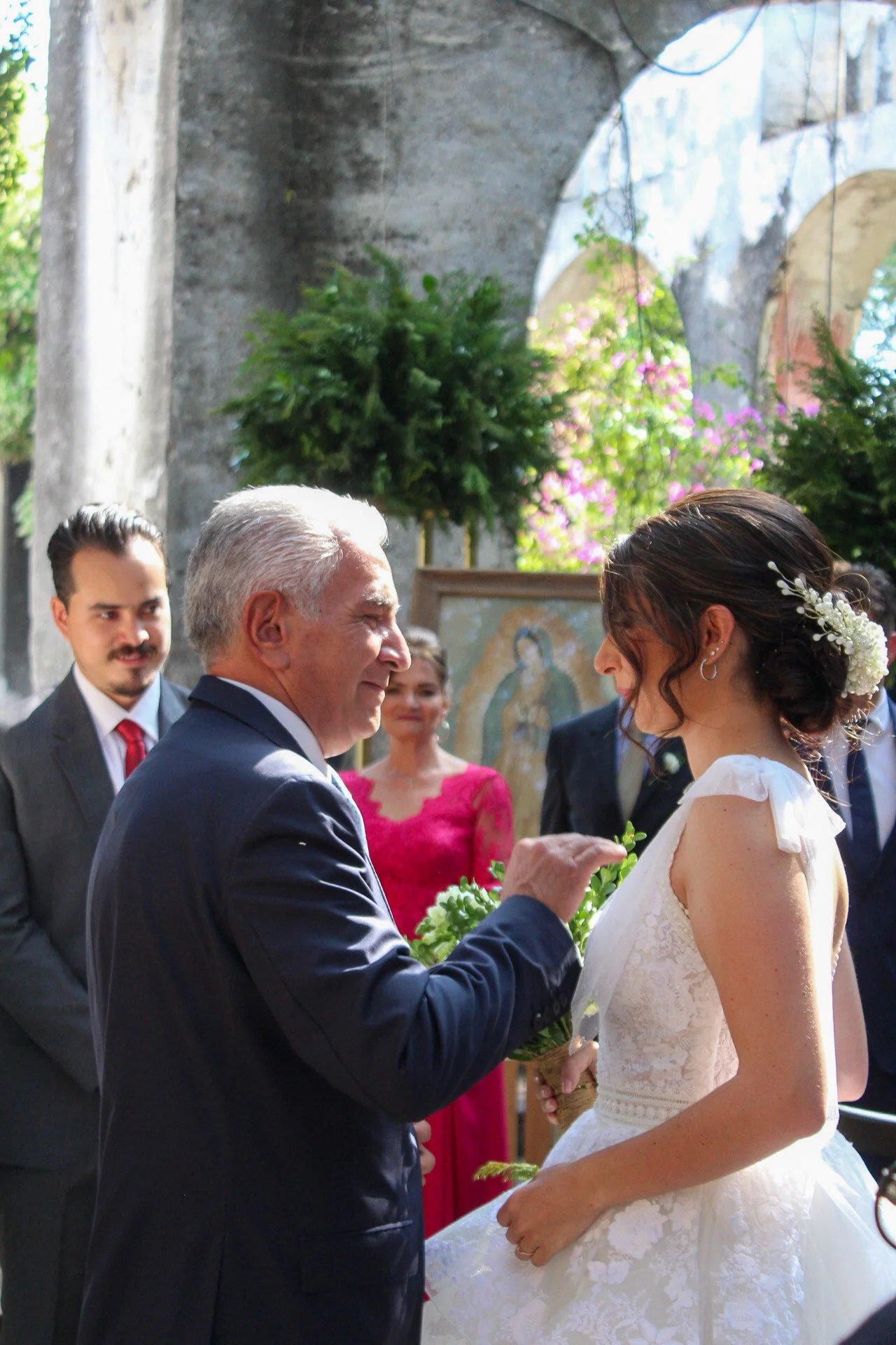 Una pareja en una boda, el hombre mayor y la mujer joven están en un intercambio de votos o promesas, rodeados de familiares y amigos en un lugar con arcos y vegetación.