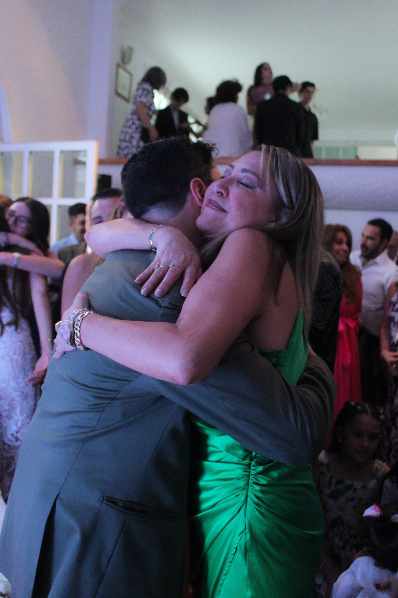 Dos personas abrazándose en una celebración, con muchas personas en el fondo, en un ambiente festivo.