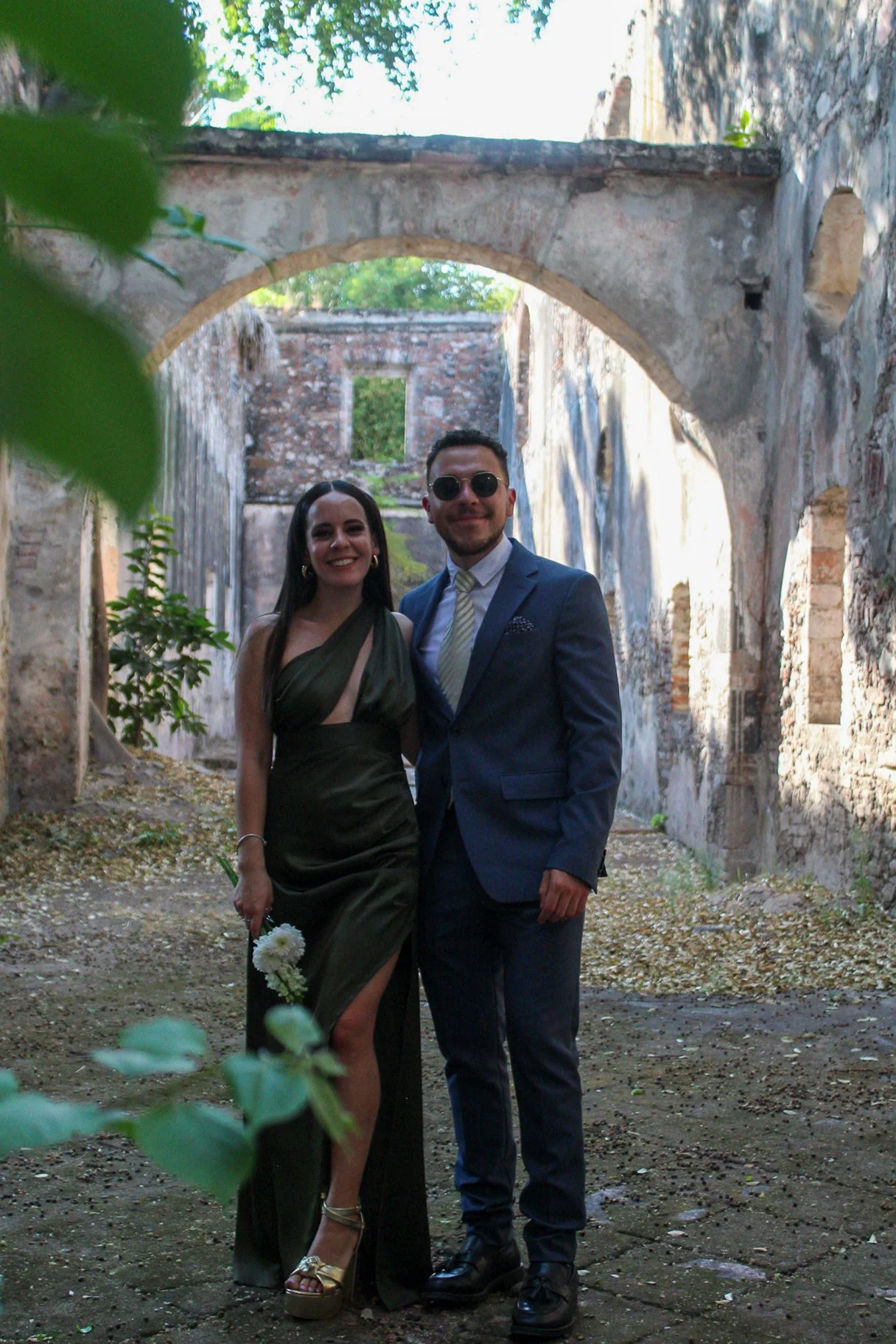 Pareja vestida con ropa formal en un lugar con ruinas antiguas y vegetación, posando juntos y sonriendo.
