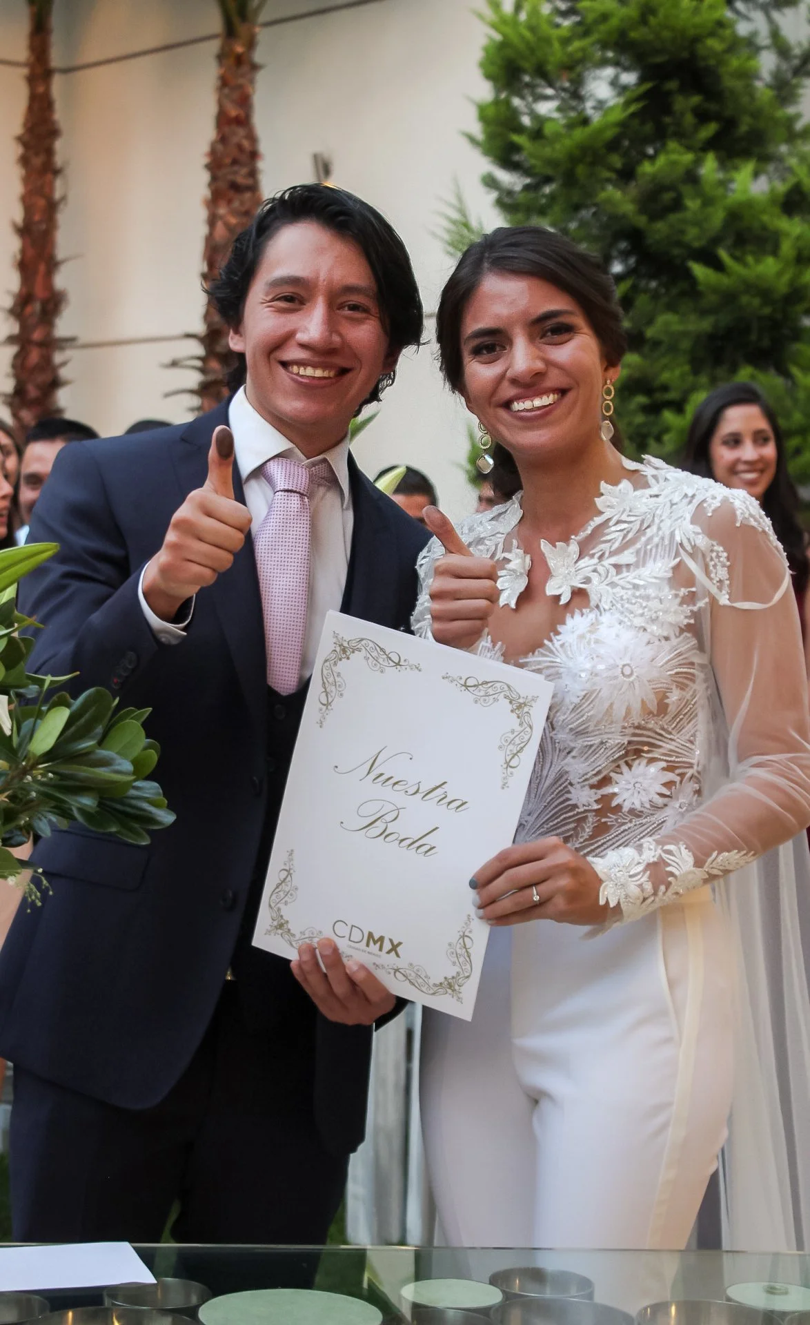 Pareja en boda mostrando un certificado que dice 'Nuestra Boda' en CDMX, ambos sonriendo y haciendo señal de 'pulgar arriba'.
