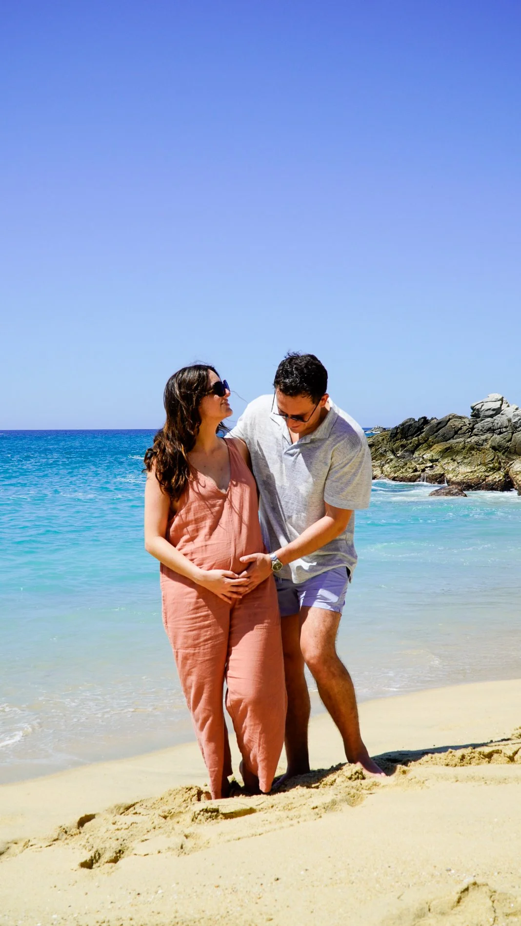 Pareja en la playa, la mujer embarazada y el hombre se abrazan y sonríen, con el mar y rocas de fondo.