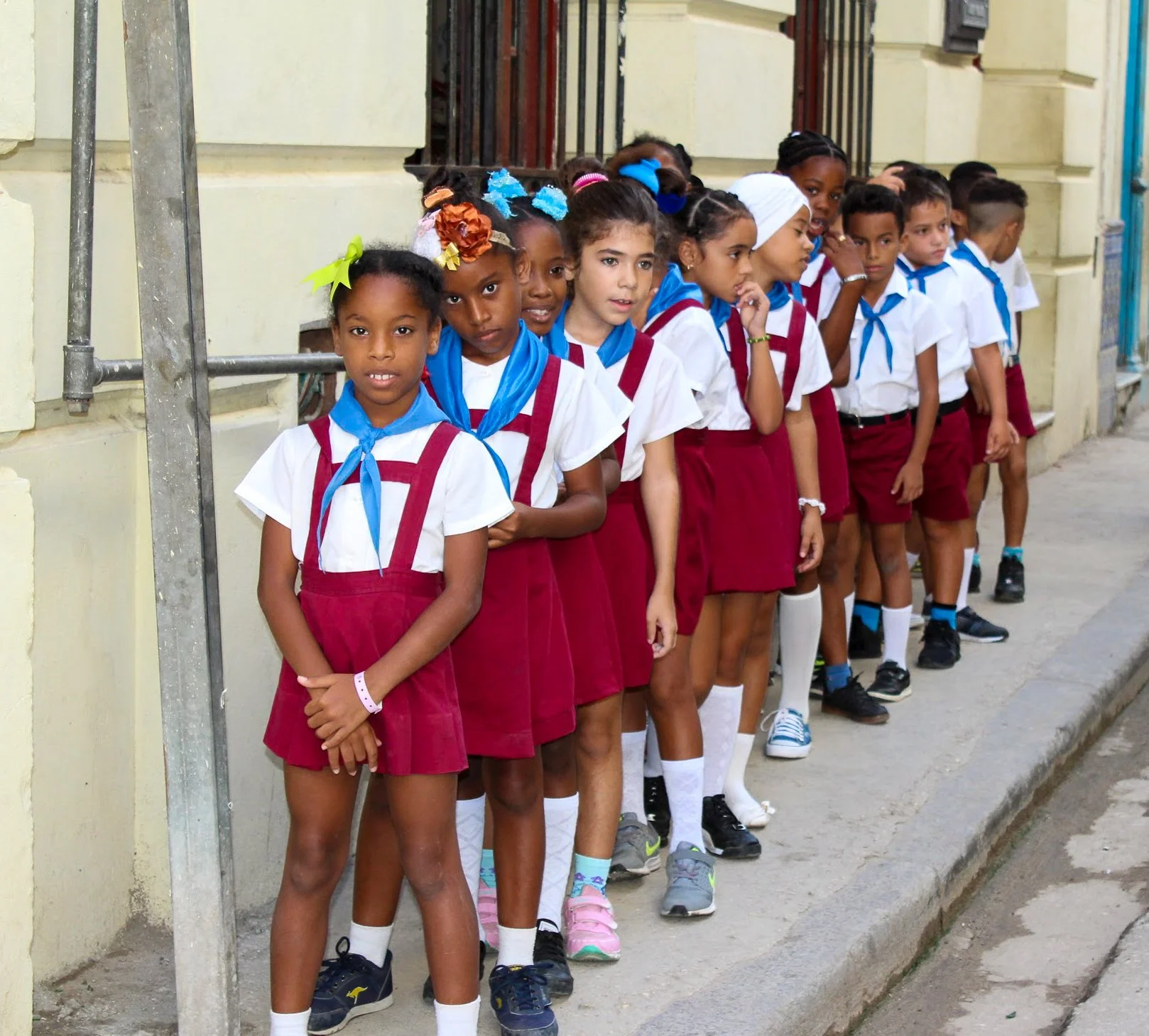 Niños y niñas en fila en uniforme escolar, algunos con diferentes accesorios de color y estilos, en la acera.