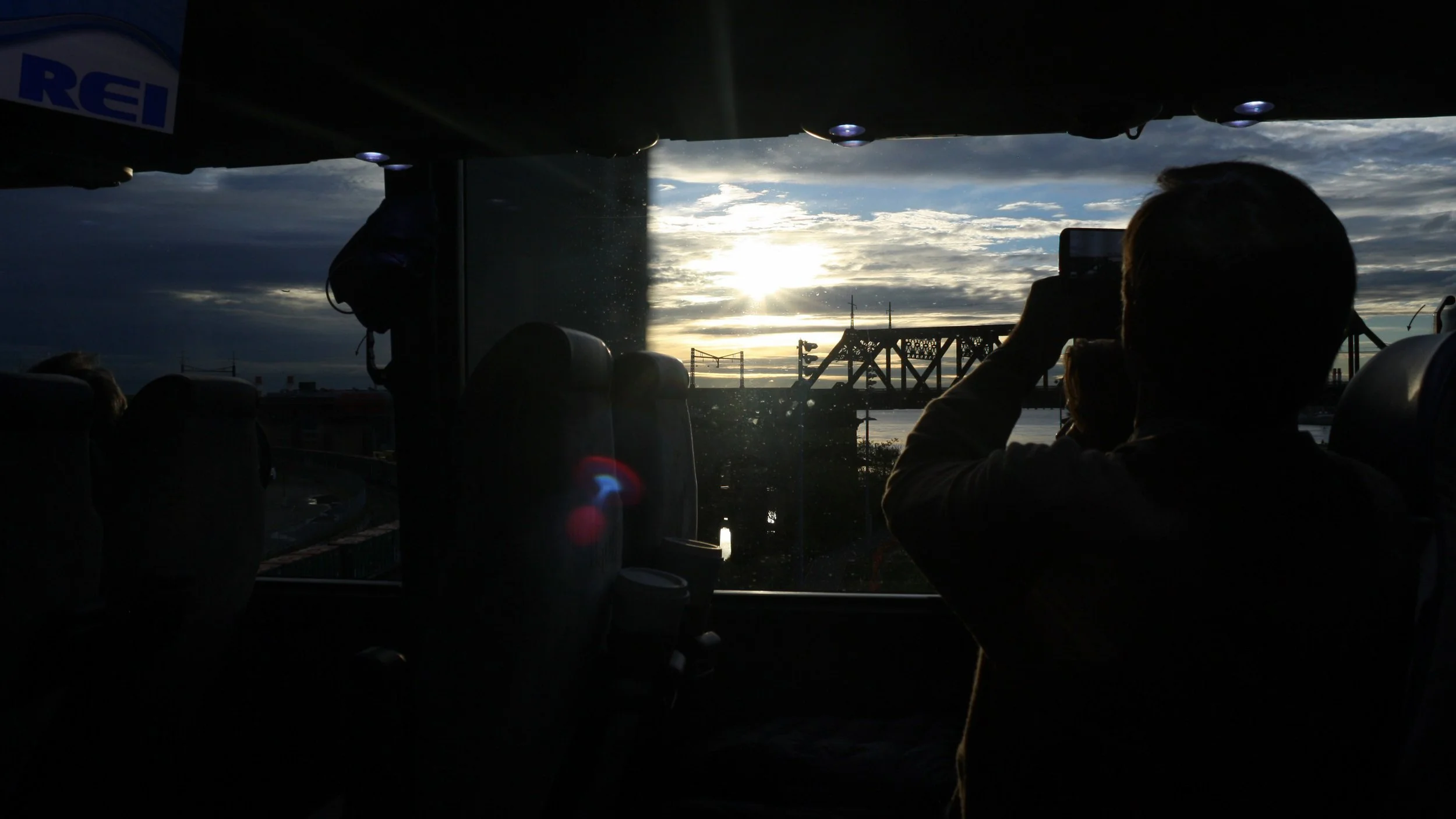 Interior de un autobús con una persona tomando una foto del atardecer a través de la ventana.