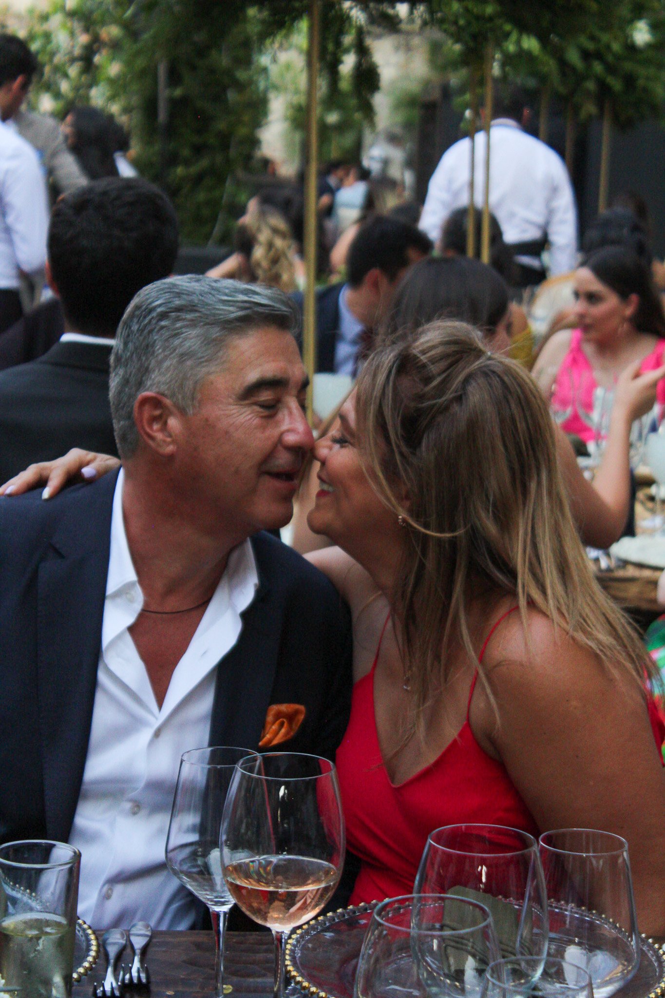 Pareja en una celebración social, se están sonriendo y tocando las narices, rodeados de otras personas en un ambiente al aire libre con muchas copas de vino y comida sobre la mesa.