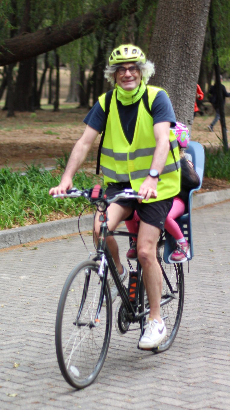 Persona mayor con gafas y casco andando en bicicleta en un parque, lleva chaleco reflectante y está sonriente.