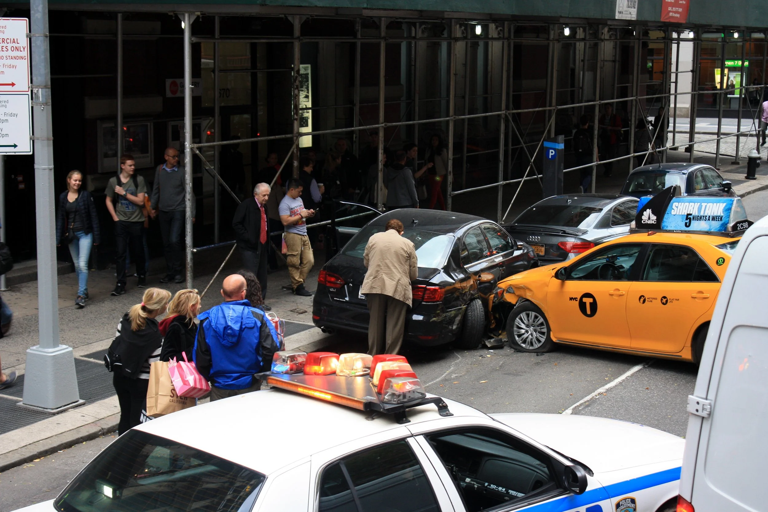 Un accidente de coche en una ciudad con varias personas alrededor, algunos observando y otros en movimiento. Se observan varios vehículos, incluido un taxi amarillo y un coche negro, con daños visibles, y una patrulla policial en la escena.