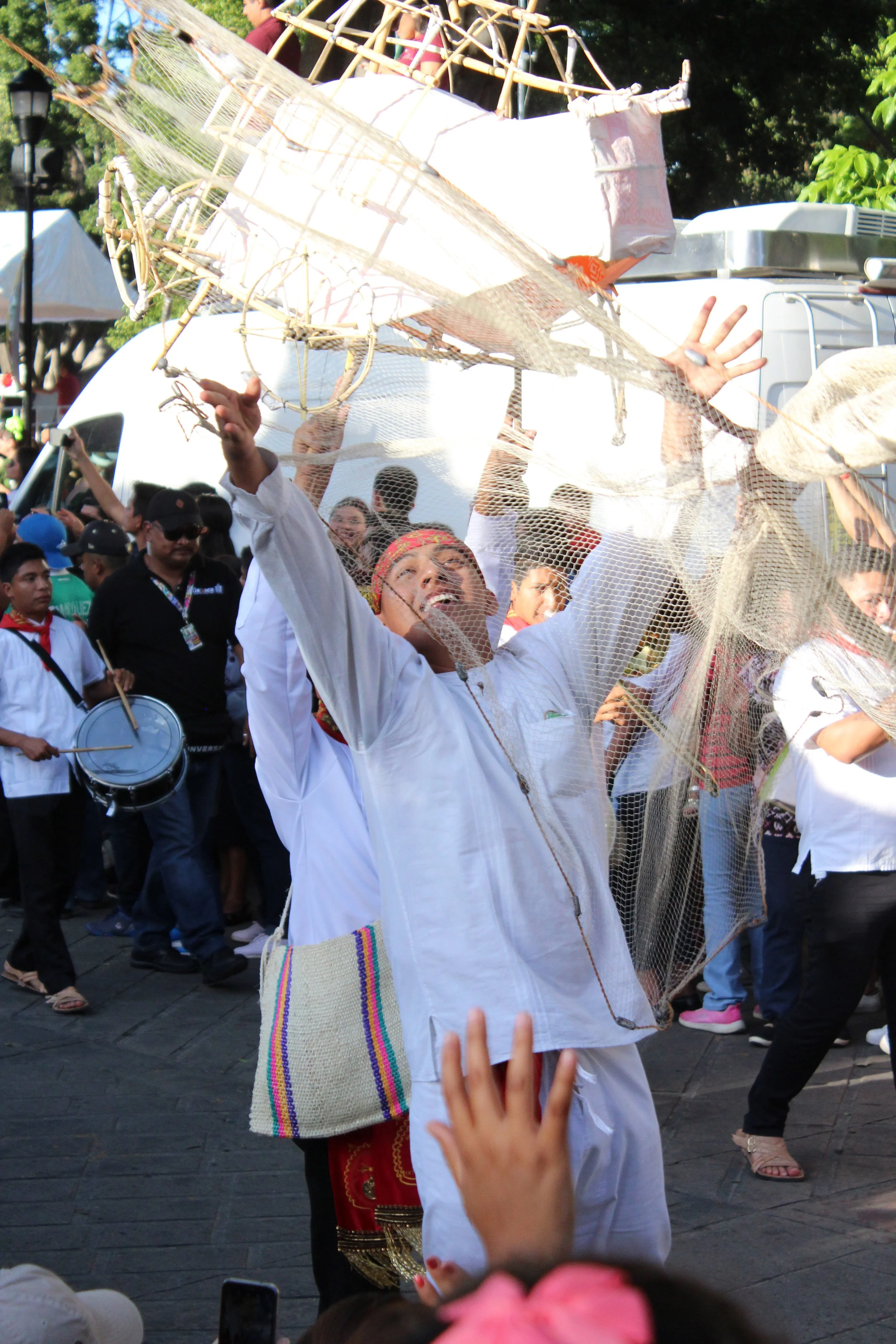 Persona participando en una festividad, levantando y sosteniendo un objeto decorativo elaborado con malla y madera, rodeada de otras personas en un evento al aire libre.