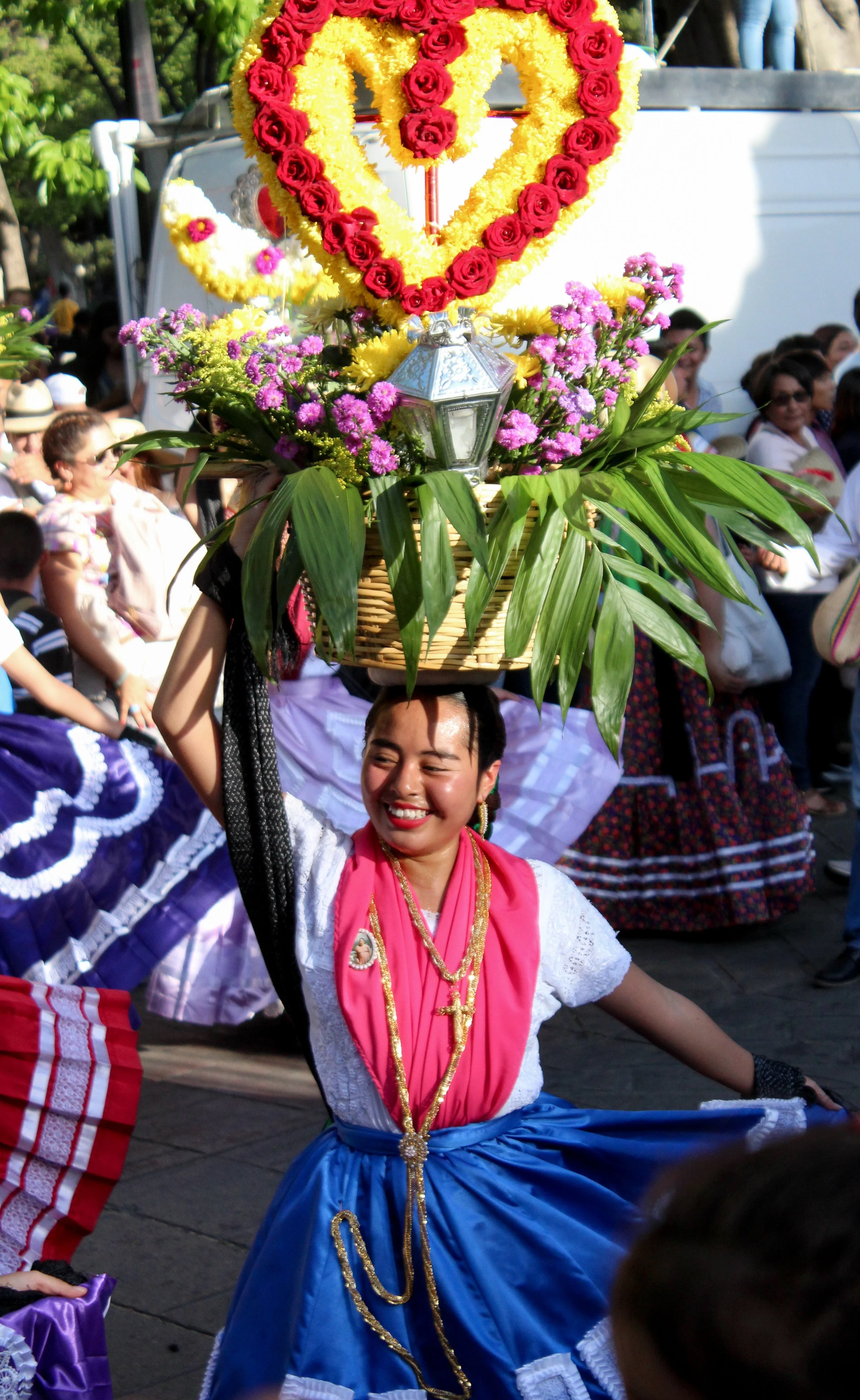 Mujer vestida con traje tradicional, bailando en un desfile cultural, llevando un canasto con flores sobre la cabeza.