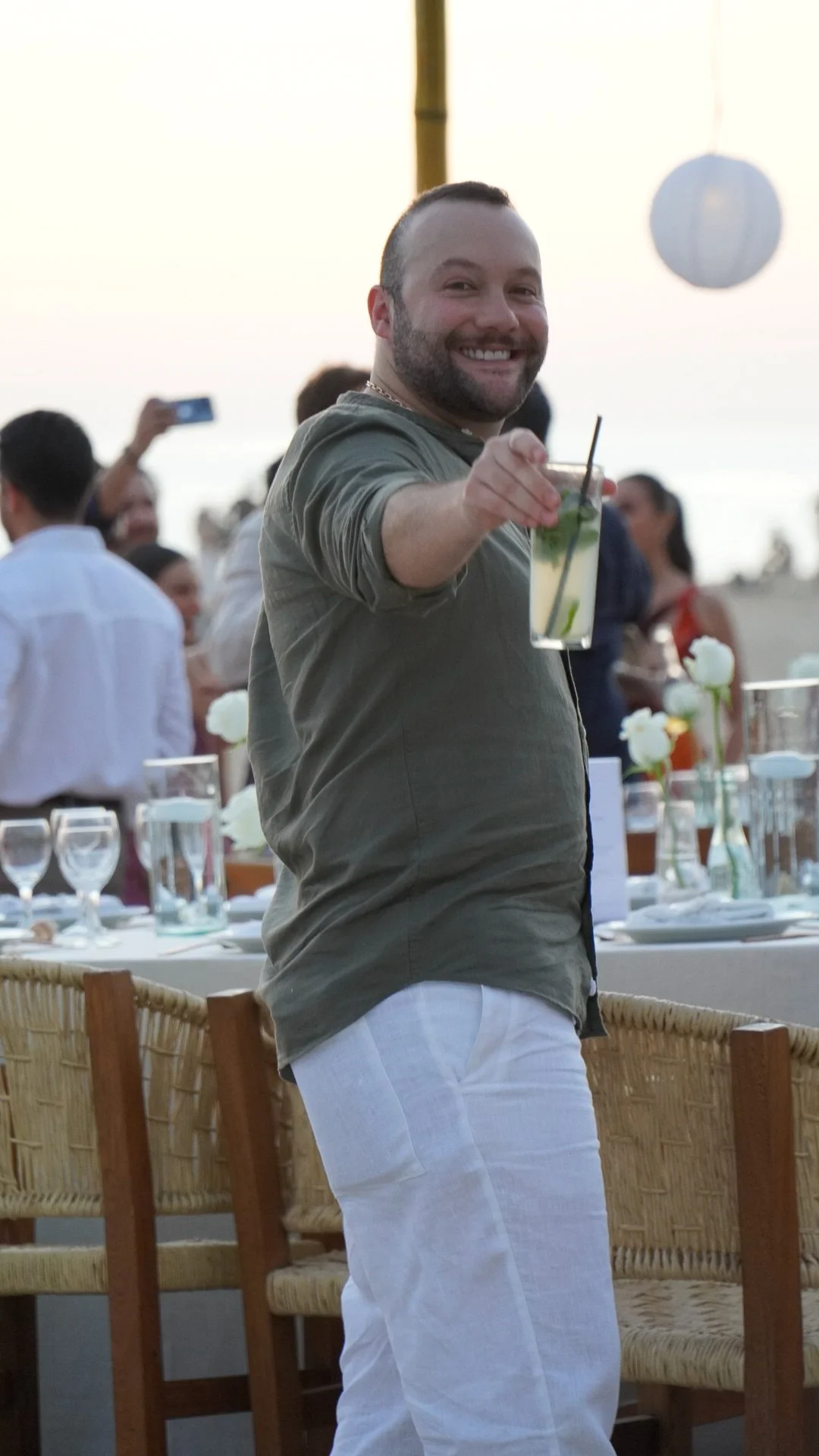 Hombre sonriendo en una fiesta en la playa, sostiene una bebida en un vaso alto con rodaja de limón y menta, fondo con mesas decoradas y personas tomando fotos.