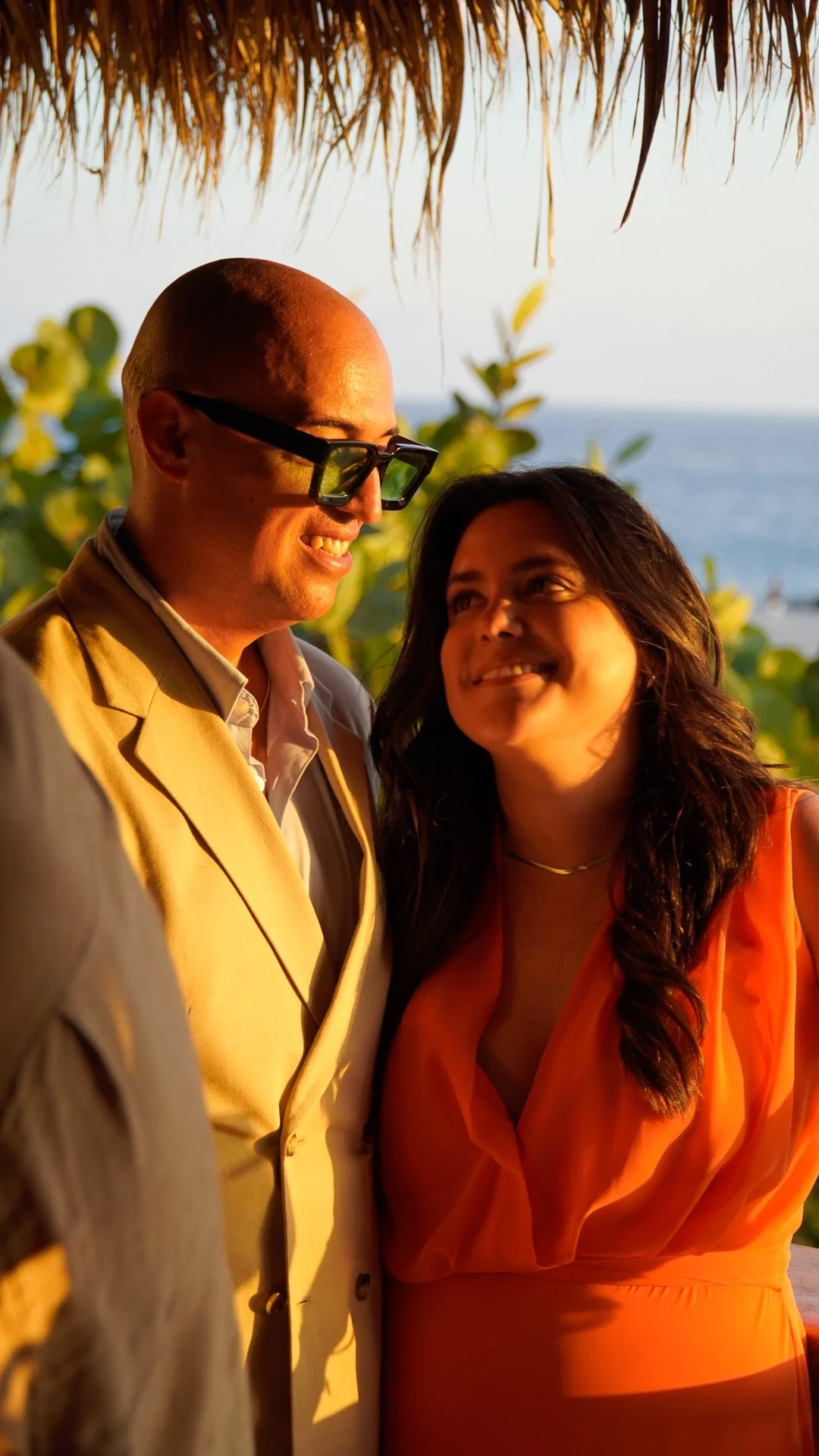 Una pareja sonriendo en un entorno tropical al atardecer, con vegetación y el mar al fondo.