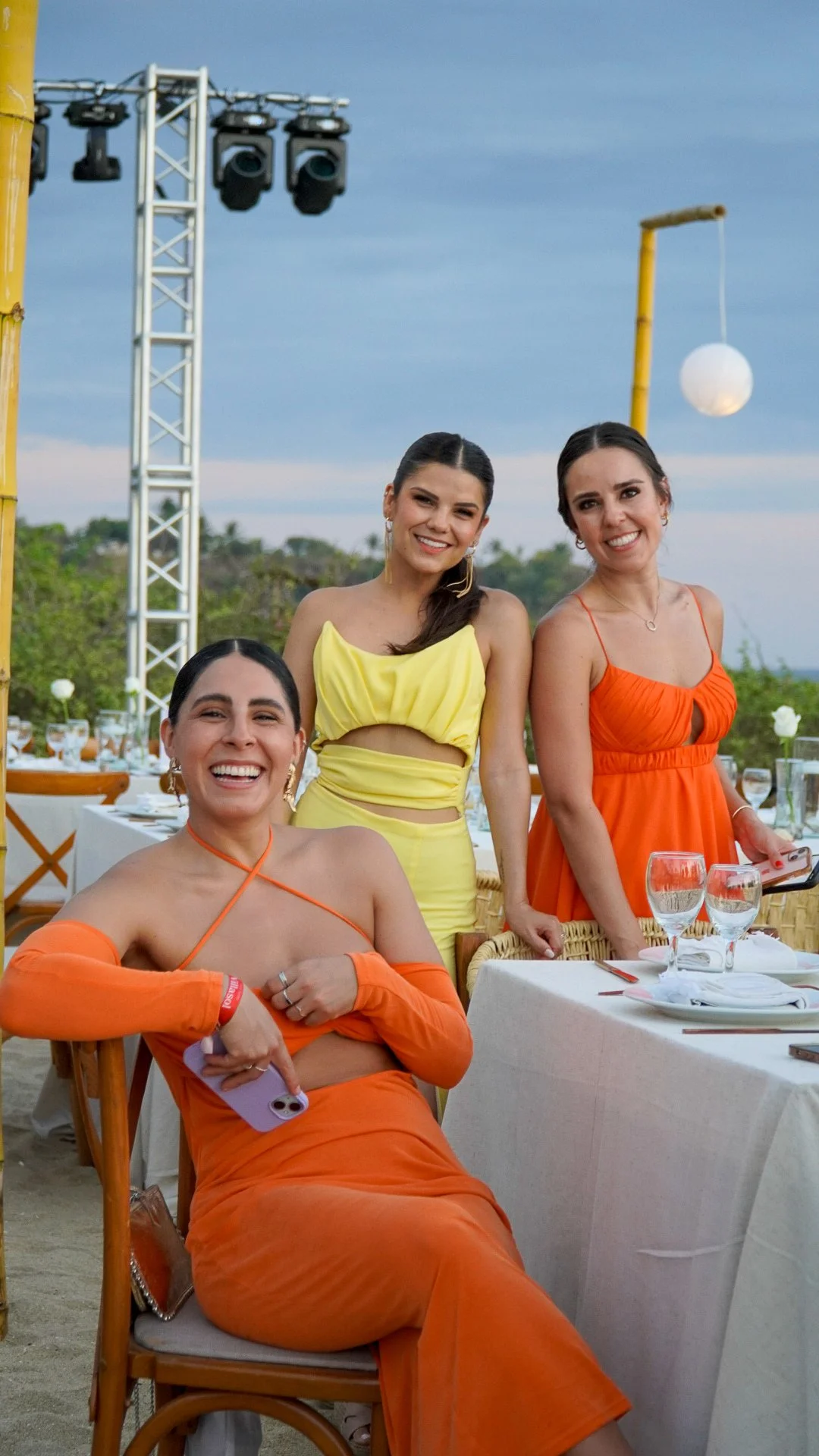 Tres mujeres sonriendo en una fiesta al aire libre con decoración colorida y luces, en una mesa con copas y platos, durante el atardecer.