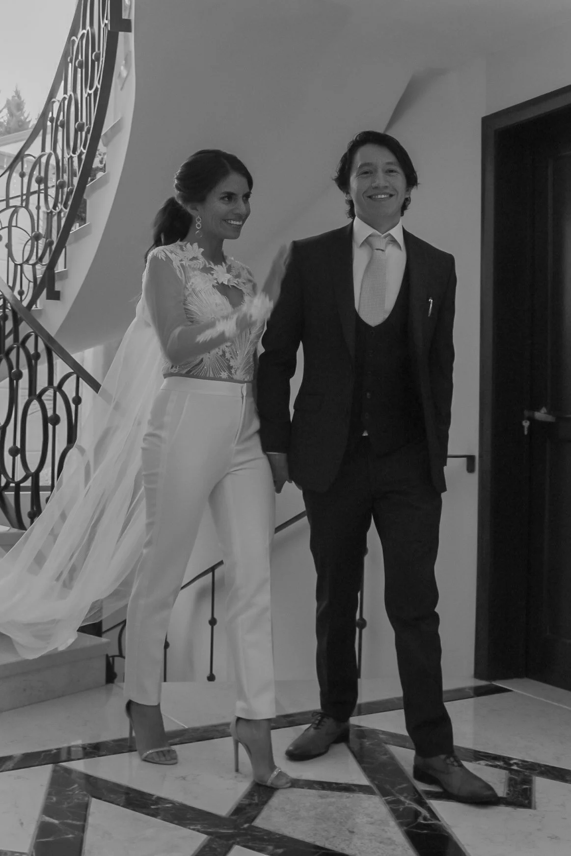 Pareja de novios en una boda, la mujer lleva un vestido de novia y el hombre un traje formal, en un interior con escaleras y suelo de mosaico.