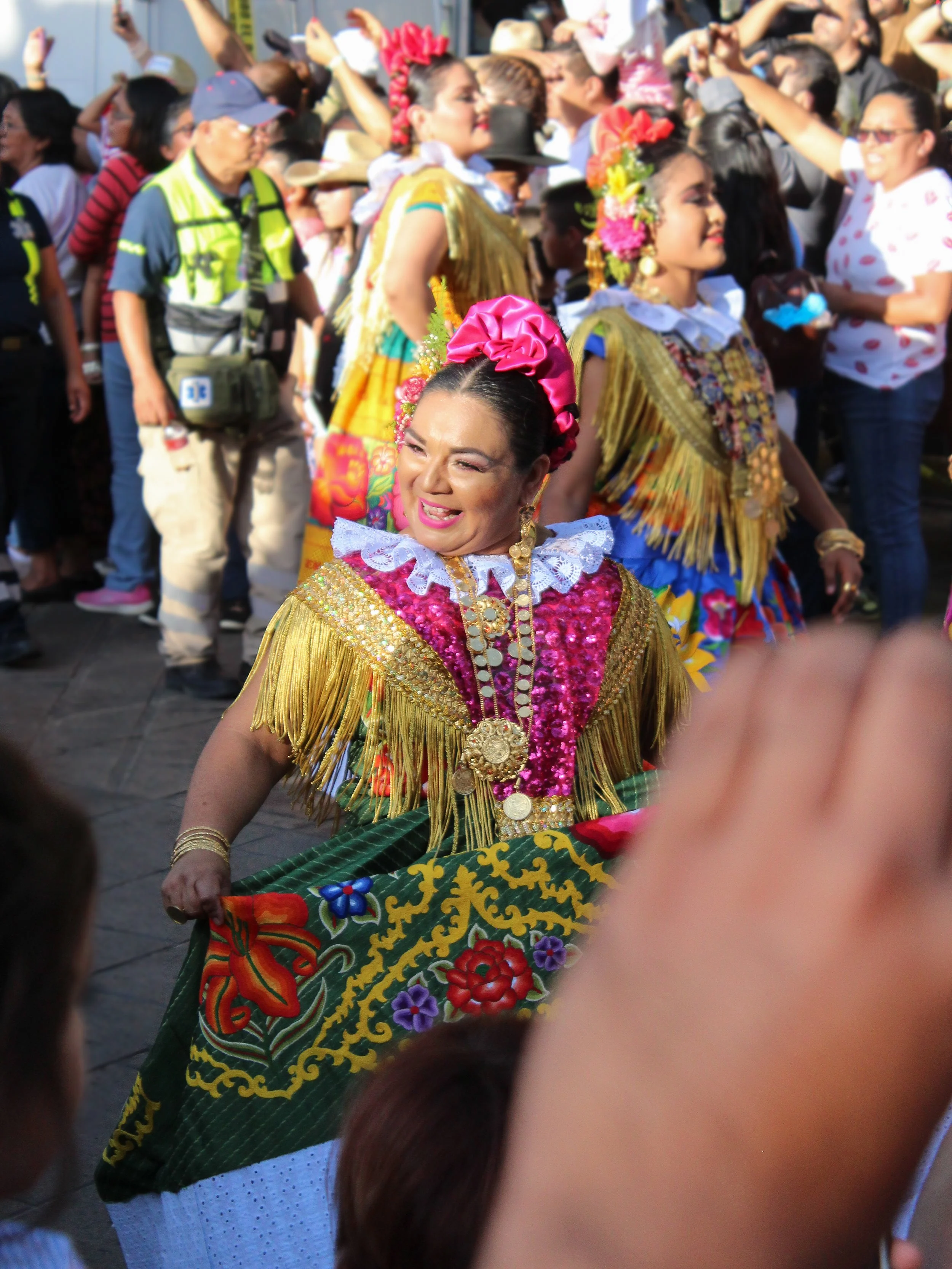 Mujer en vestimenta tradicional de celebración, bailando en un desfile, rodeada de otras personas también vestidas con trajes típicos y espectadores en el fondo.