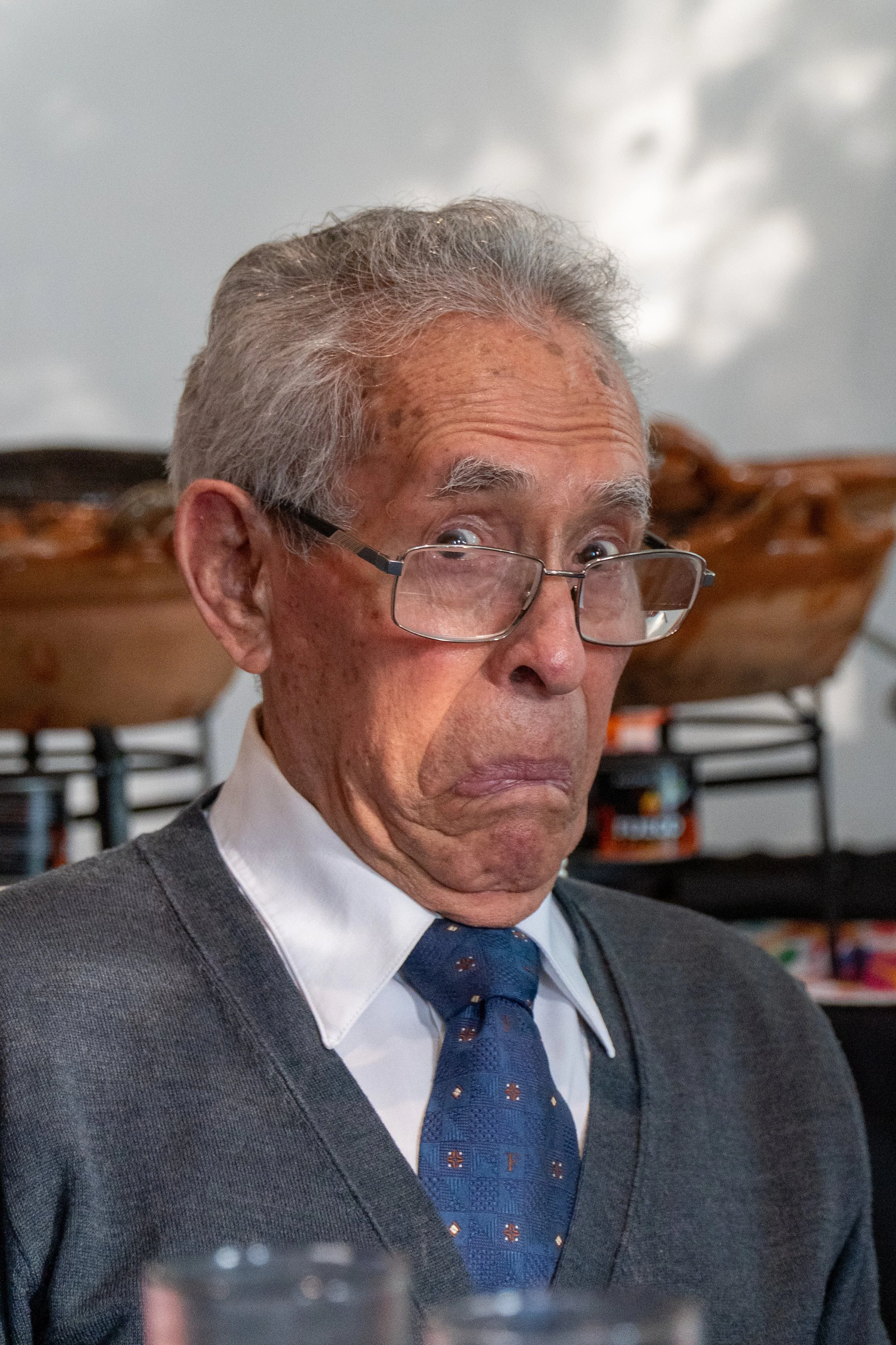 Un hombre mayor con gafas, expresión de sorpresa o disgusto, lleva una camisa blanca, corbata azul y suéter gris. Fondo con platos de comida en un evento social.