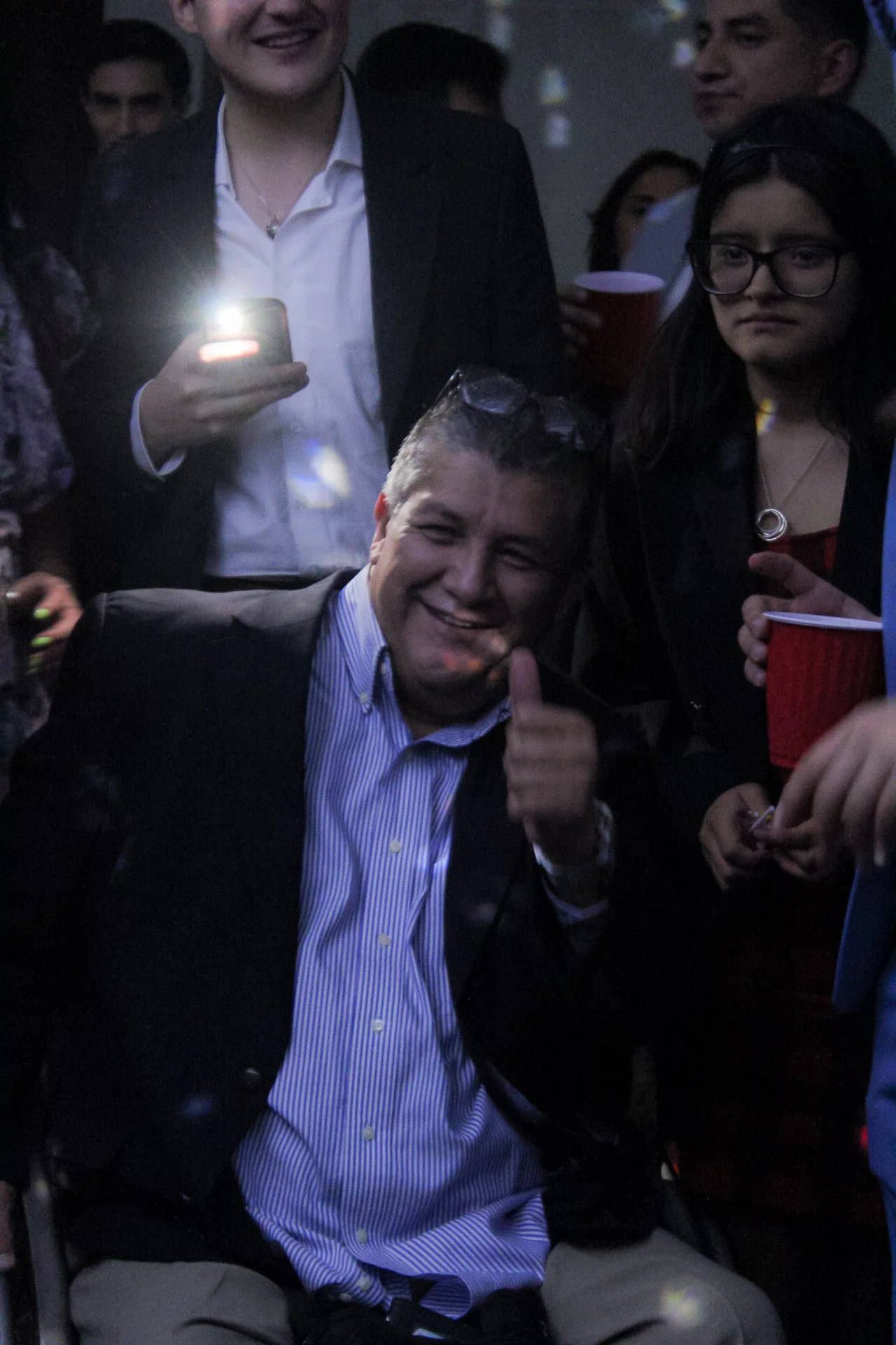 Un grupo de personas en una celebración o reunión social, con un hombre en primer plano sonriendo y haciendo un pulgar hacia arriba.