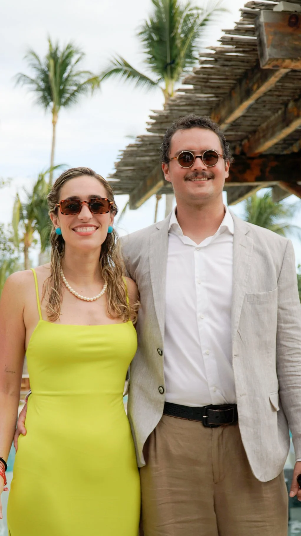 Pareja joven vestida elegantemente en un entorno tropical con palmeras y estructura de madera.