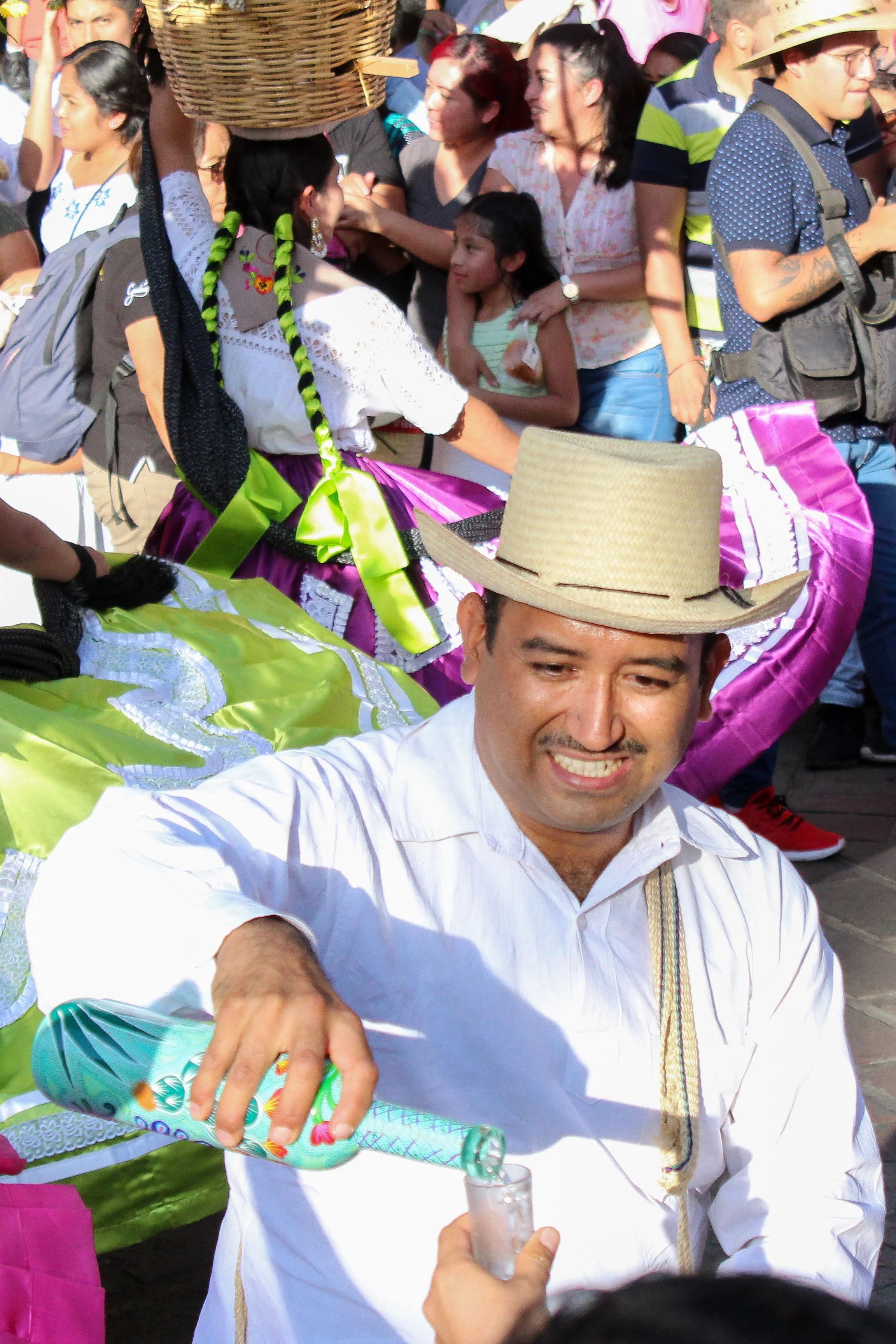 Hombre con sombrero de paja, sonriente, bebiendo una bebida durante una celebración con una multitud de personas, algunas vestidas con trajes tradicionales y coloridos.