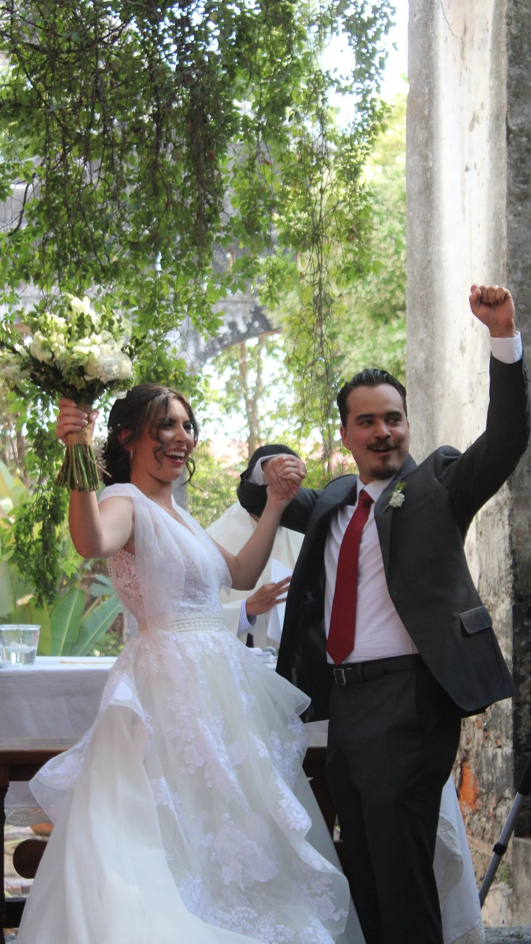 Pareja de recién casados celebrando, ella con vestido de novia y él con saco y corbata, en una ceremonia al aire libre con árboles y columnas de piedra.